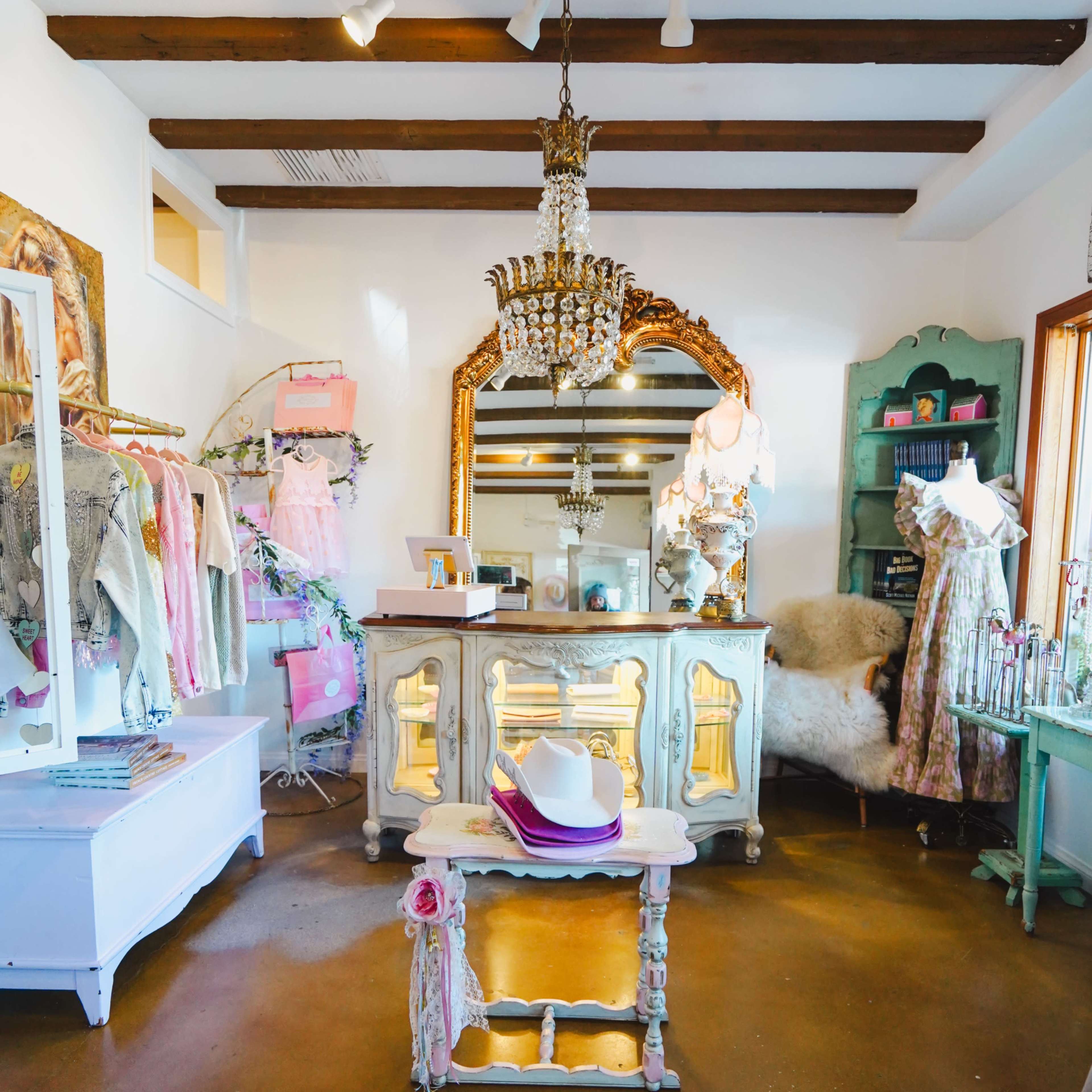 A boutique interior featuring a chandelier, a large ornate mirror, clothing displayed on racks, and a vintage-style furniture set.