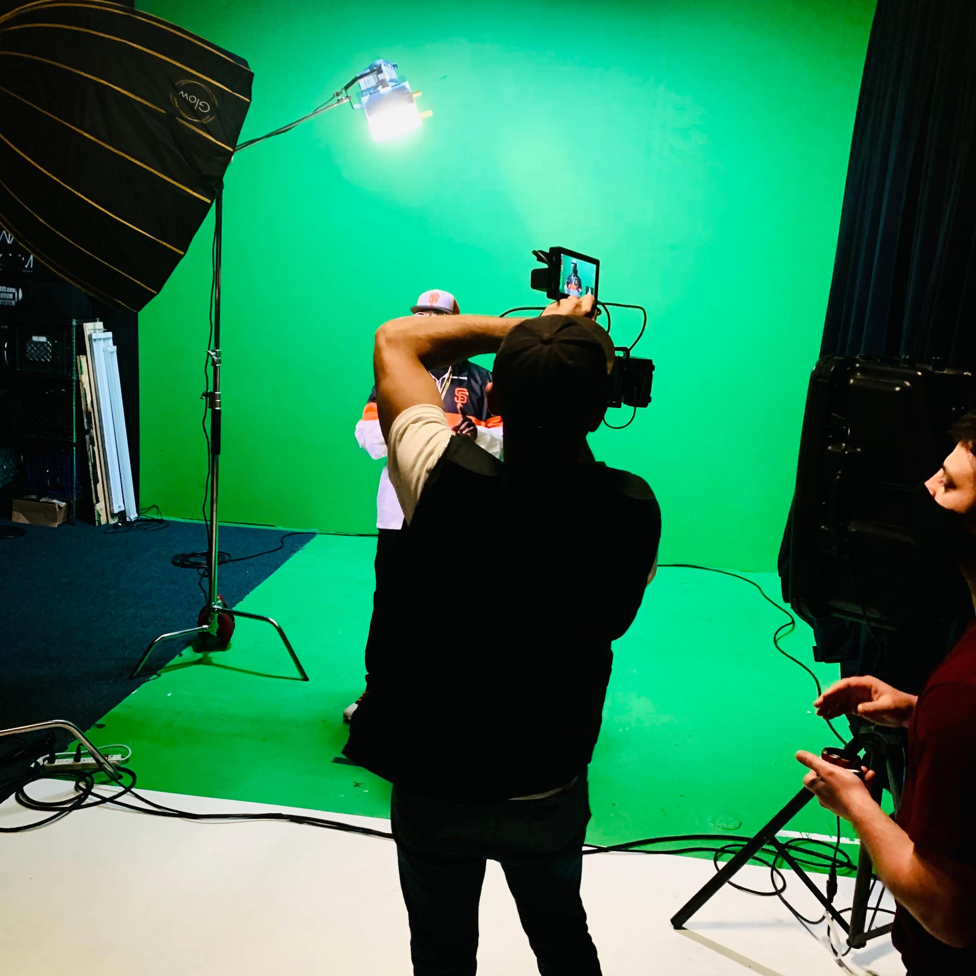 A person is filming another individual in front of a green screen, with studio lighting and equipment set up in a recording studio.