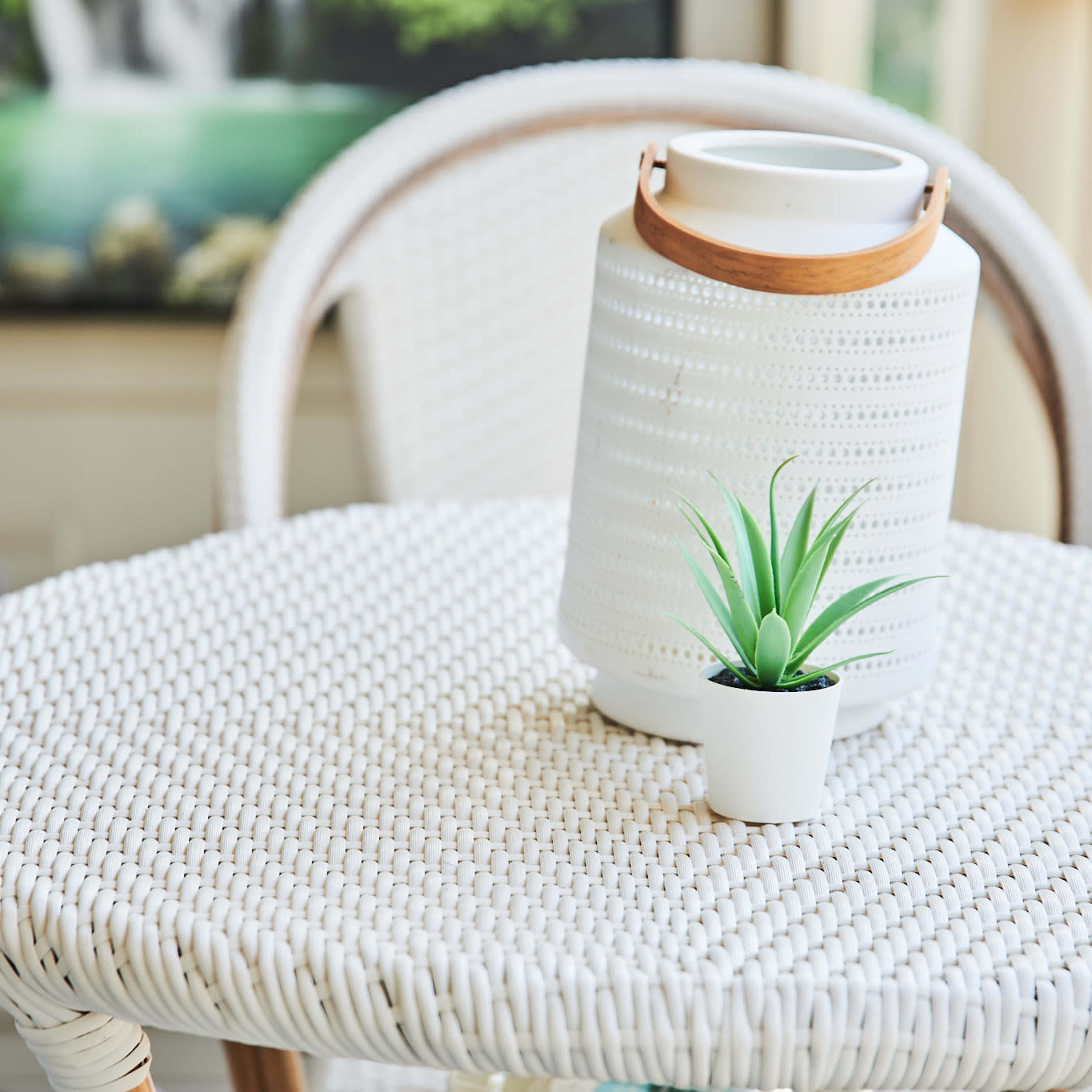 A white woven table holds a small potted plant and a lantern on a porch with a landscape painting in the background.
