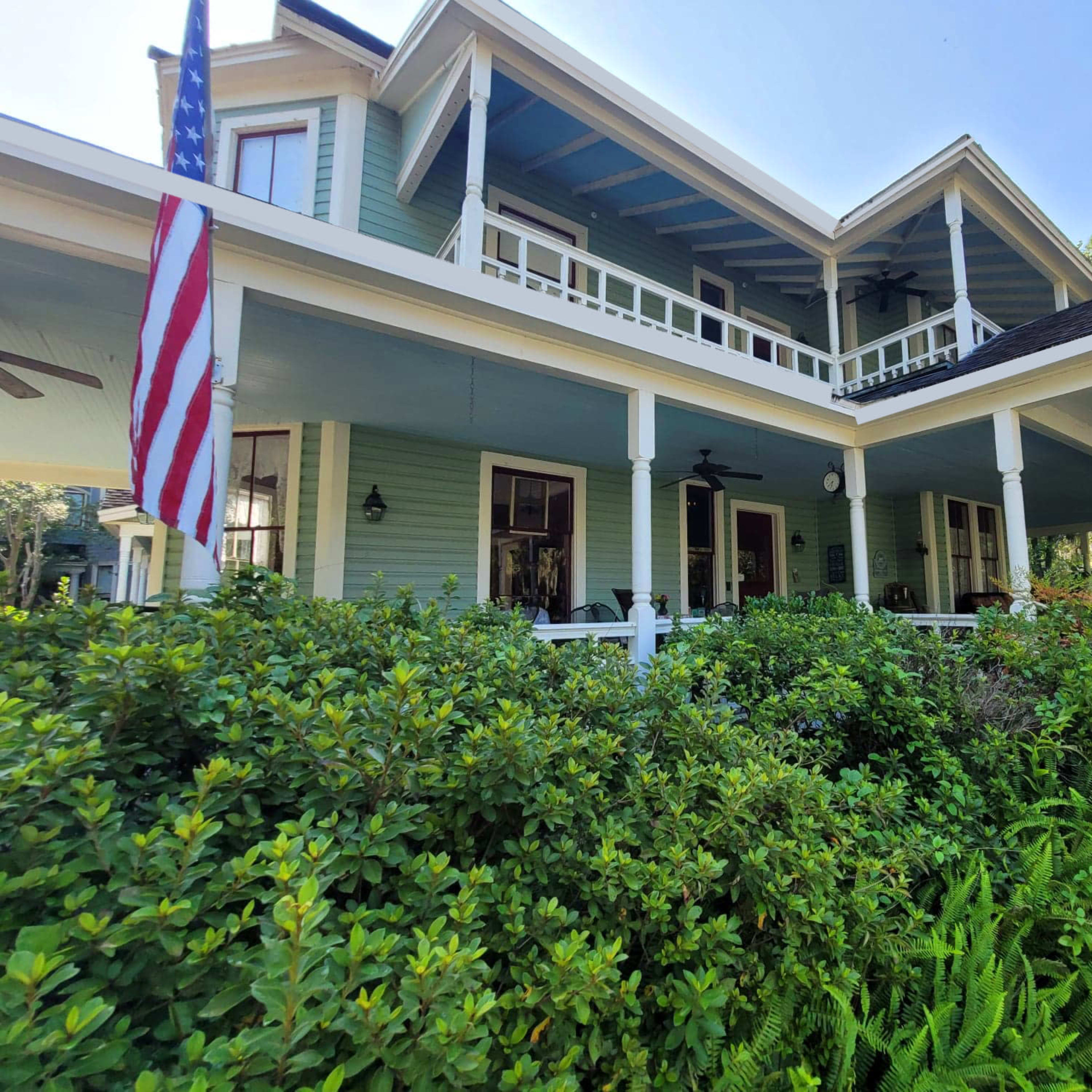 A two-story house with a wraparound porch, surrounded by greenery and featuring an American flag on the left side.
