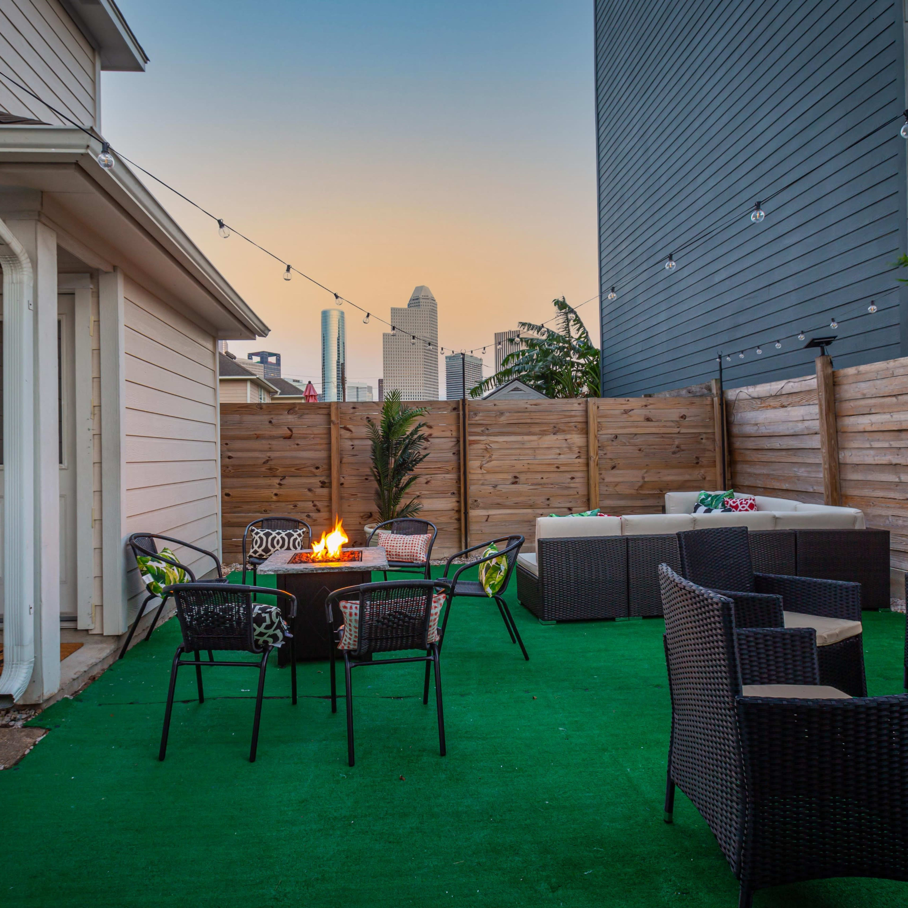 A small outdoor patio with a green rug, a fire pit surrounded by chairs, and a seating area against a wooden fence, with a skyline visible in the background.