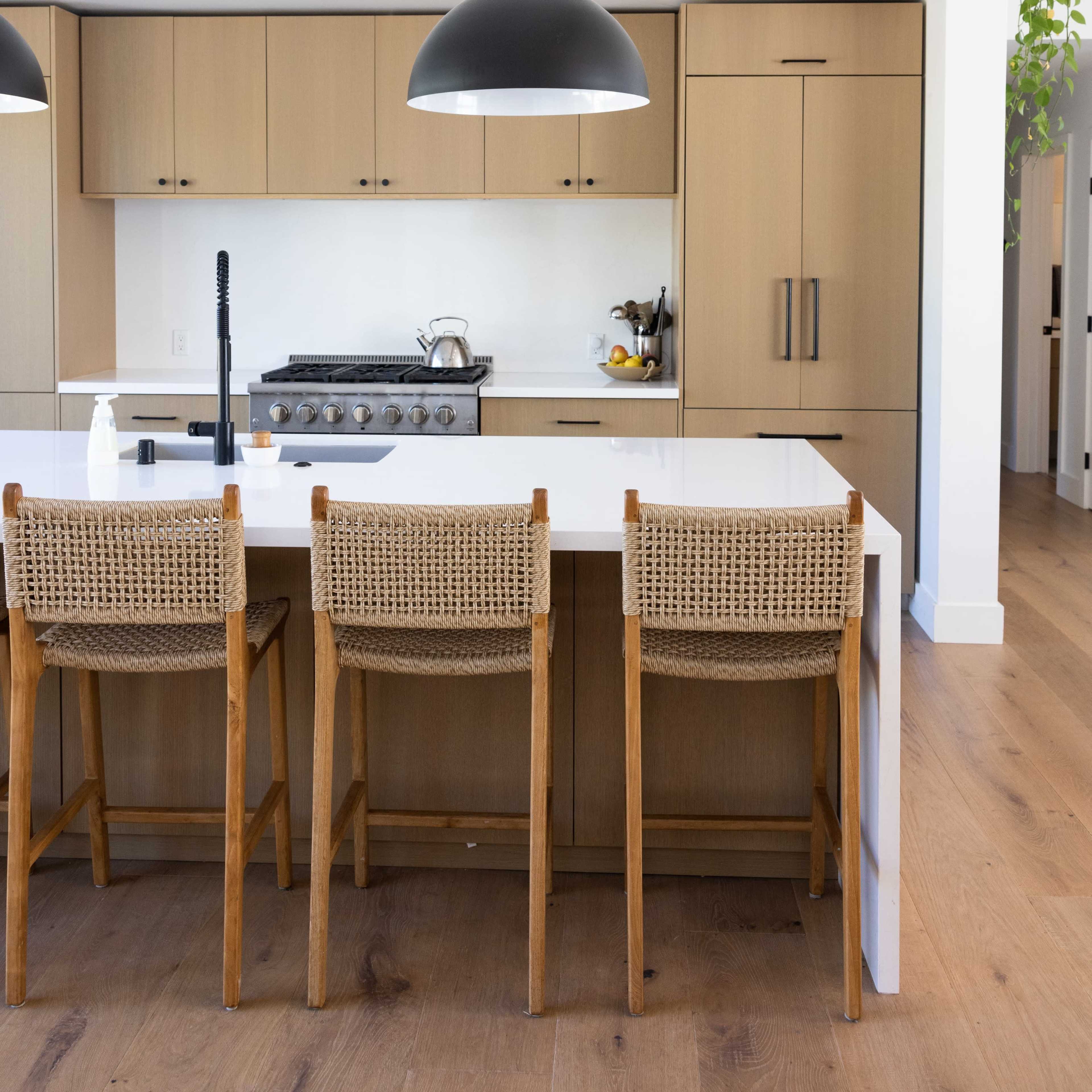 A modern kitchen features a large island with three wooden bar stools, a black pendant light overhead, and beige cabinetry.