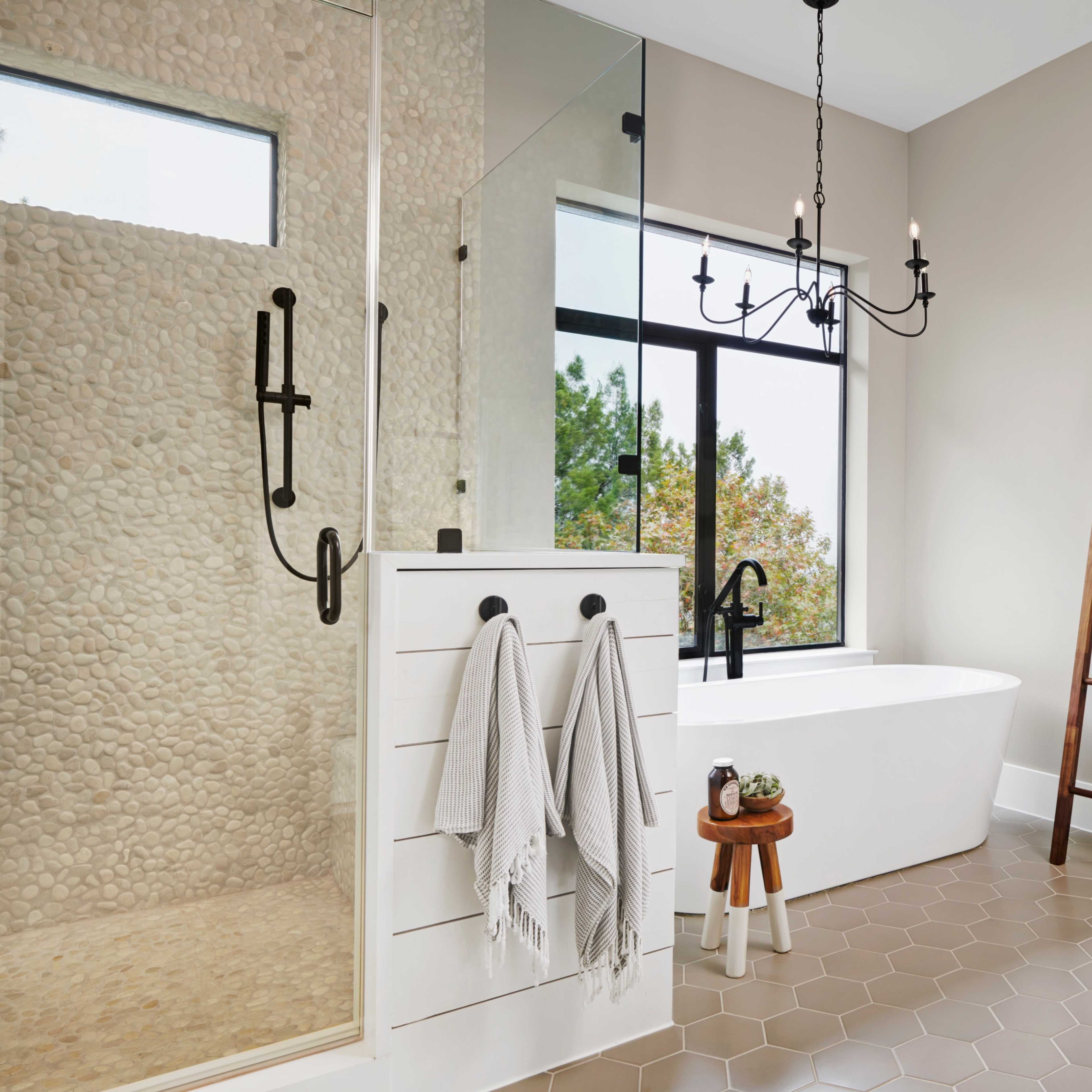 A modern bathroom with a freestanding bathtub, a glass shower enclosure, and a wooden ladder holding towels, featuring a neutral color palette and natural light from a large window.