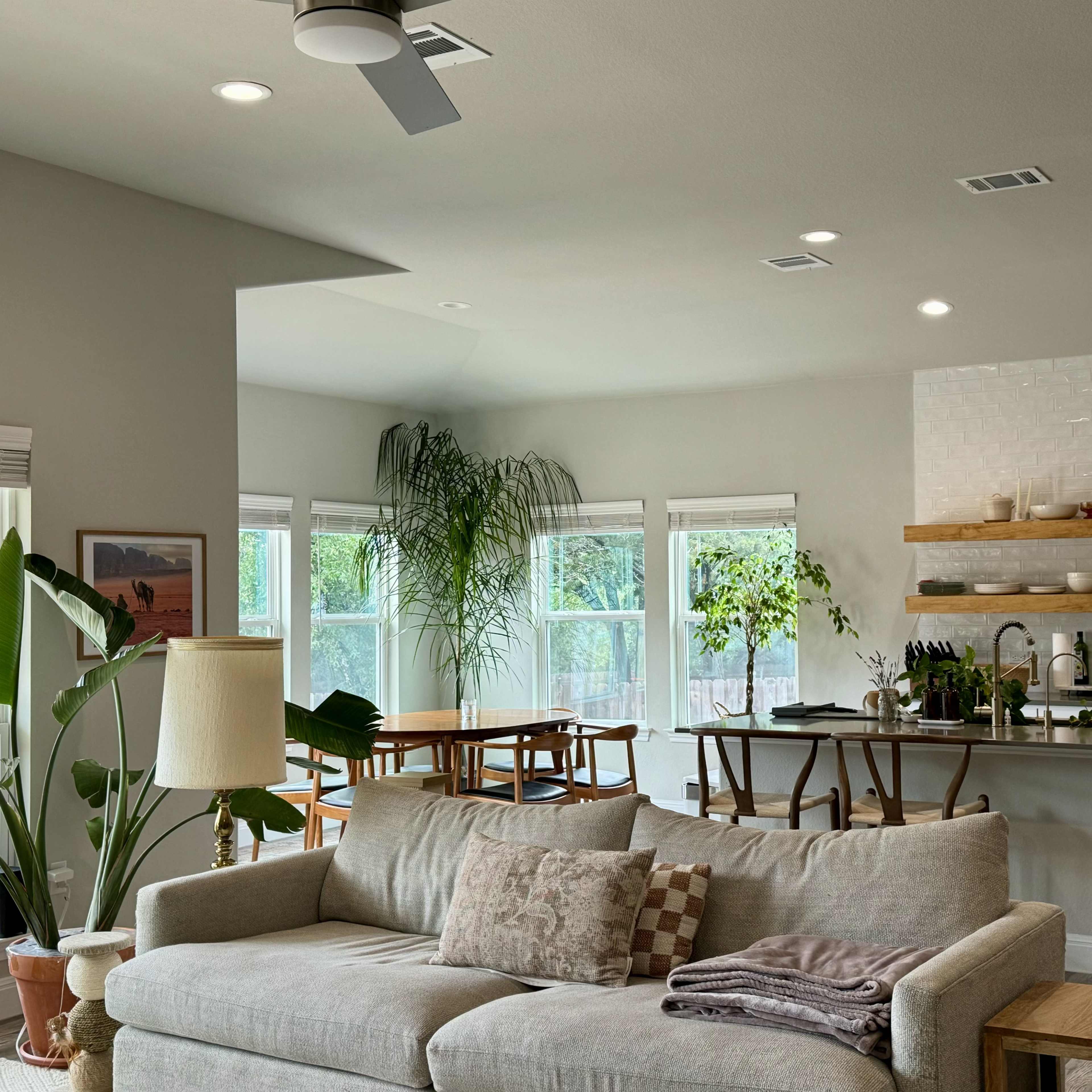 A bright, open living space featuring a light-colored sofa, a dining table surrounded by chairs, and a kitchen area with modern appliances and natural light flowing through large windows.