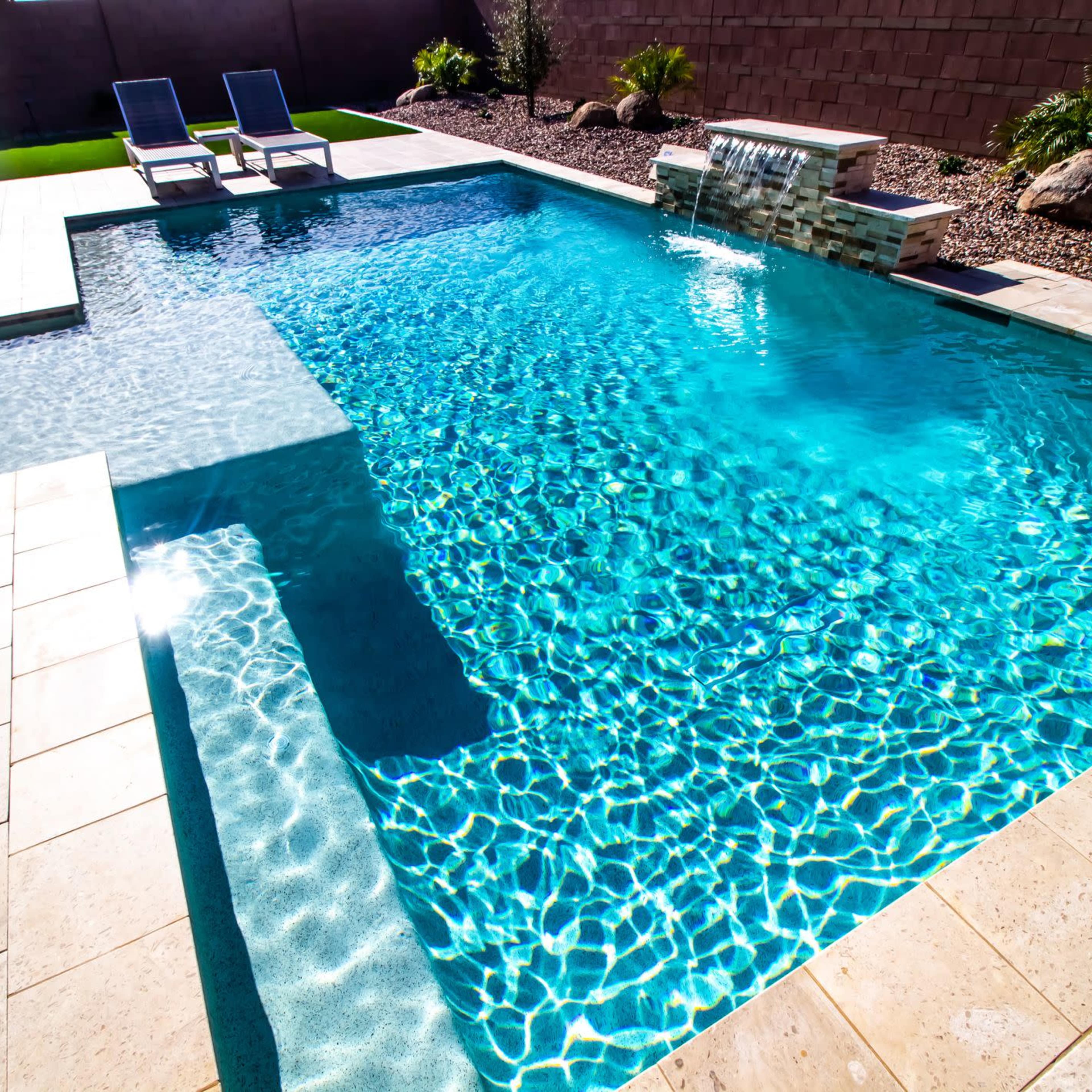 A clear, blue swimming pool with a spa section and a waterfall feature, surrounded by landscaped stones and chairs.