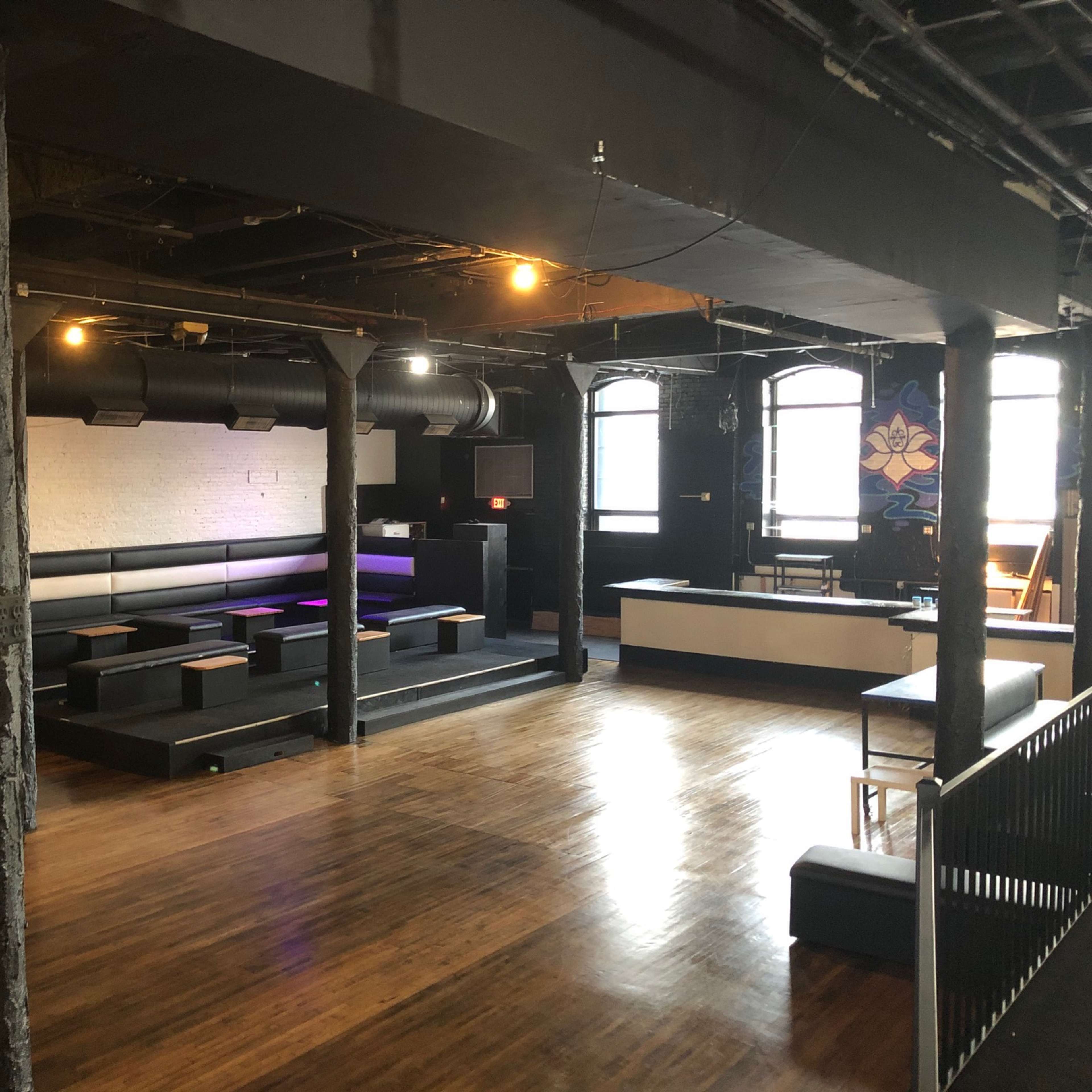 An empty interior space with wooden flooring, black seating along the walls, exposed pipes, and a bar area in the corner.