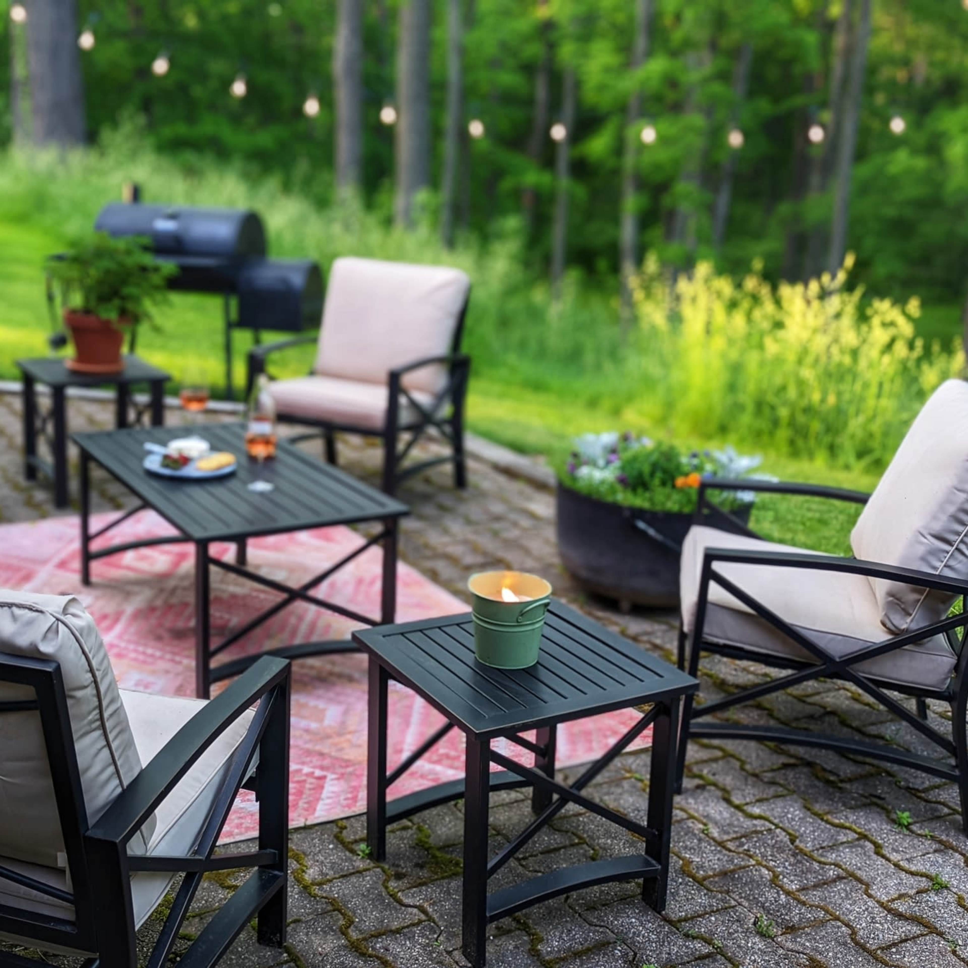 A patio area with several black metal chairs and tables arranged on a grassy lawn, surrounded by trees and string lights, featuring a small plant and refreshments on the tables.