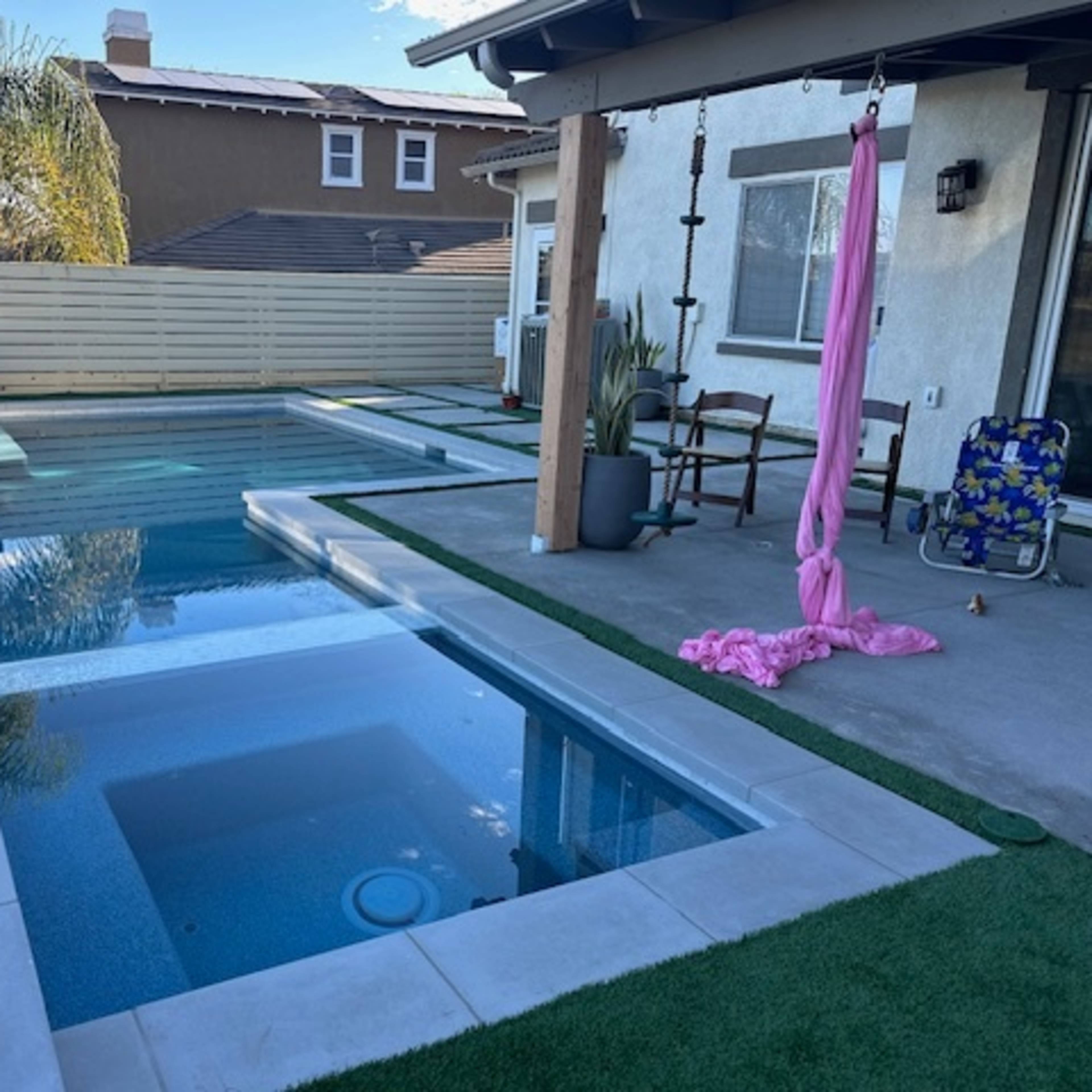 A backyard with a rectangular swimming pool and a spa, surrounded by artificial grass and a patio area with outdoor furniture.