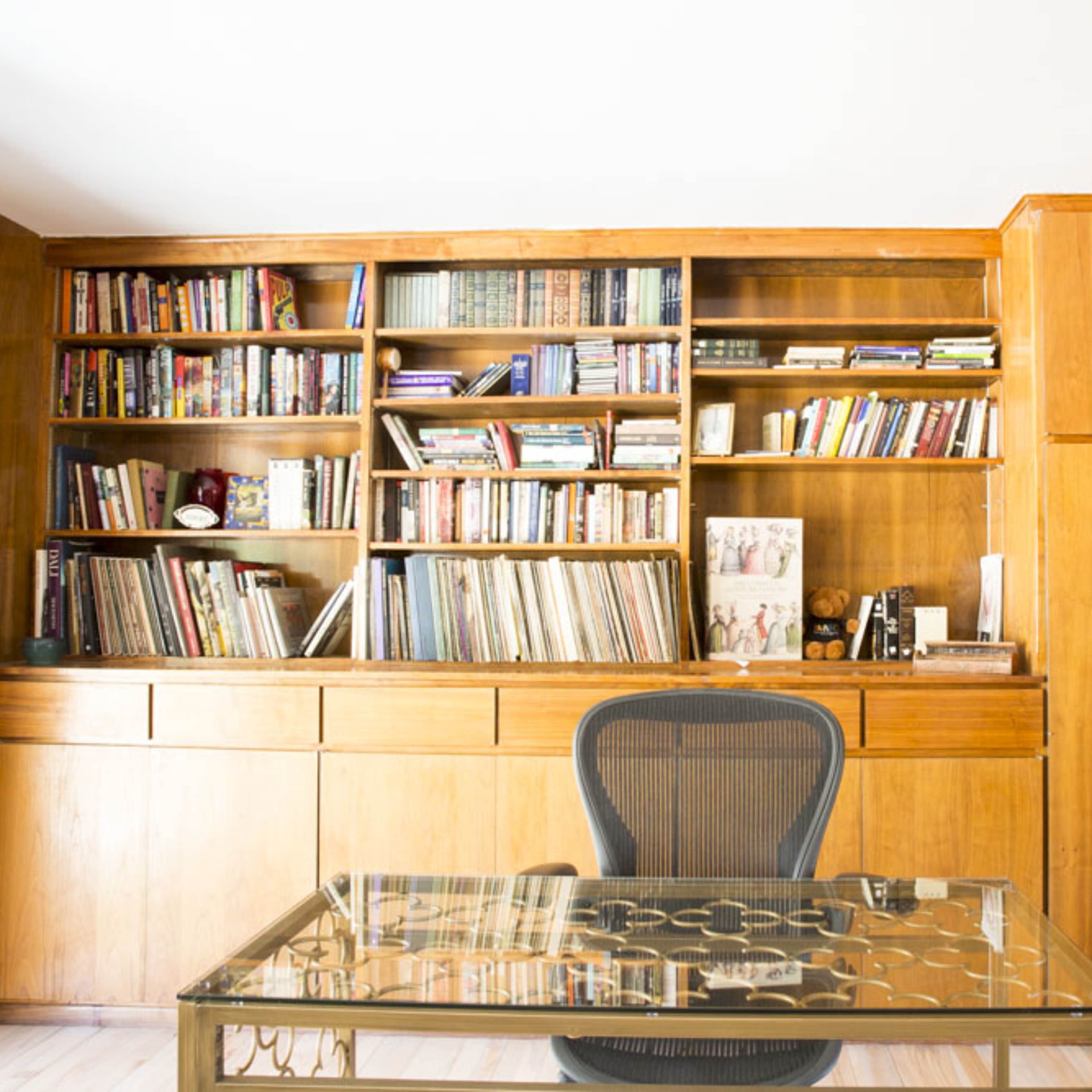 A wooden study features a large bookshelf filled with books and a glass-top desk in front of a black office chair.