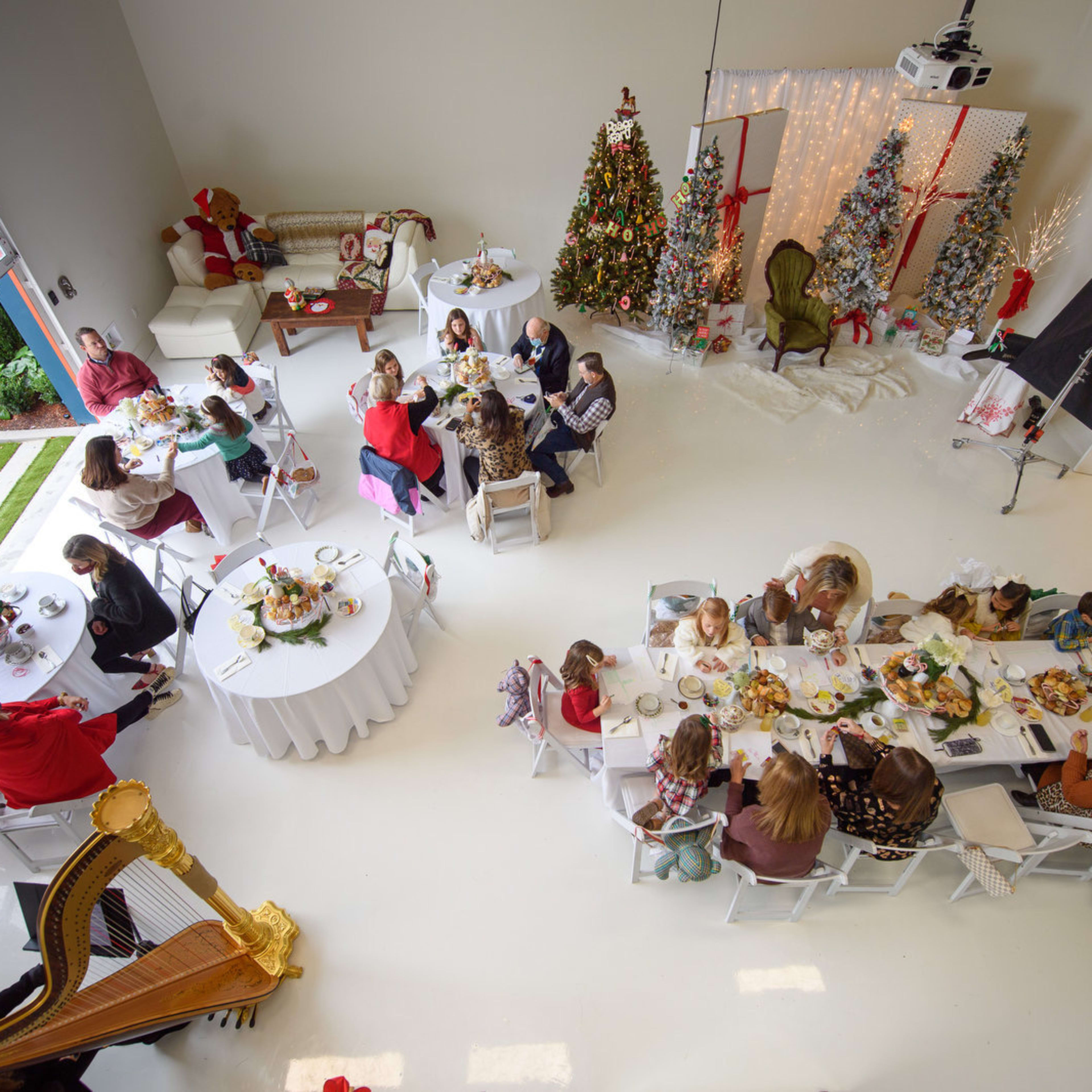 A large gathering of people is seated around tables in a spacious room decorated with Christmas trees and festive decor.