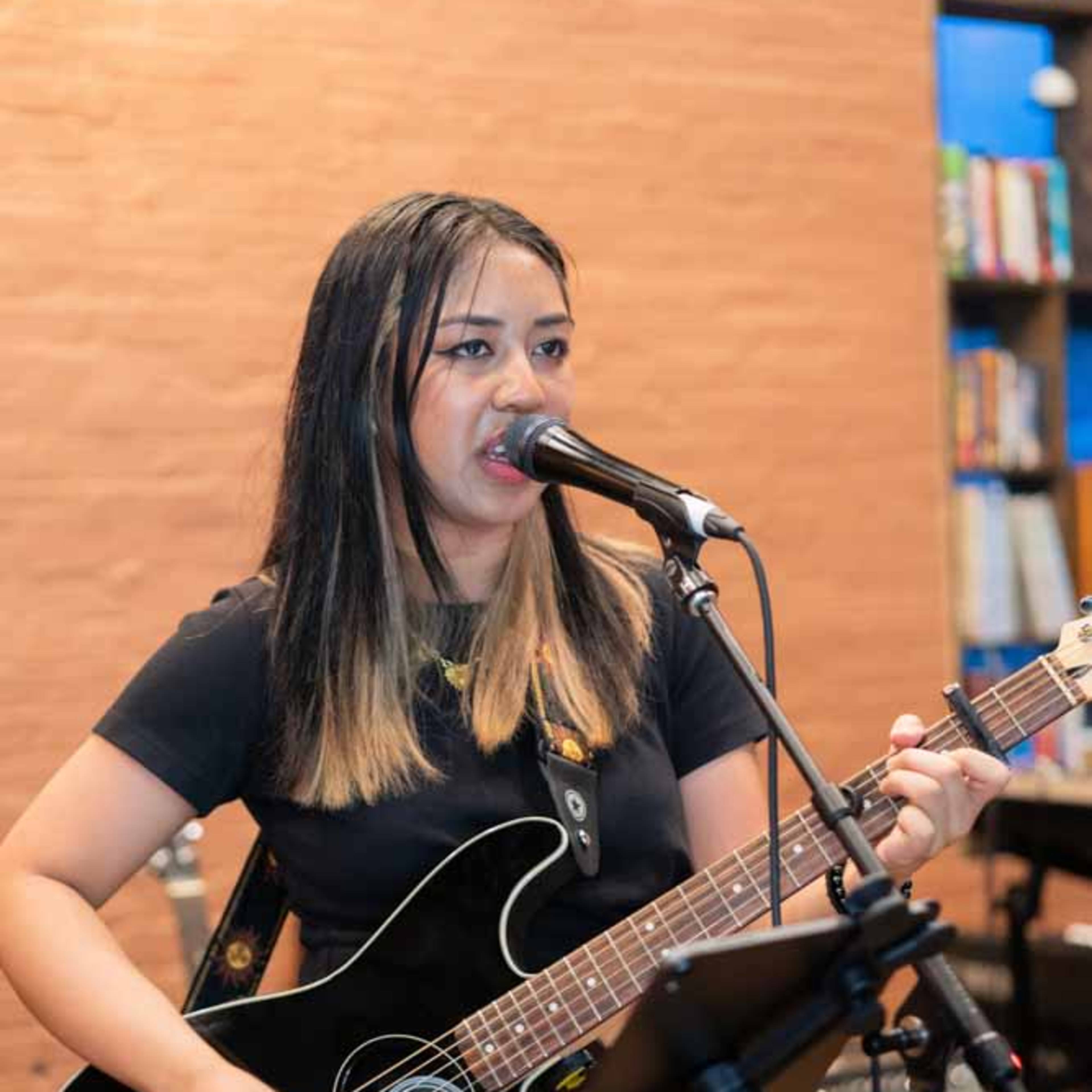 A woman sings into a microphone while playing an acoustic guitar during a performance.