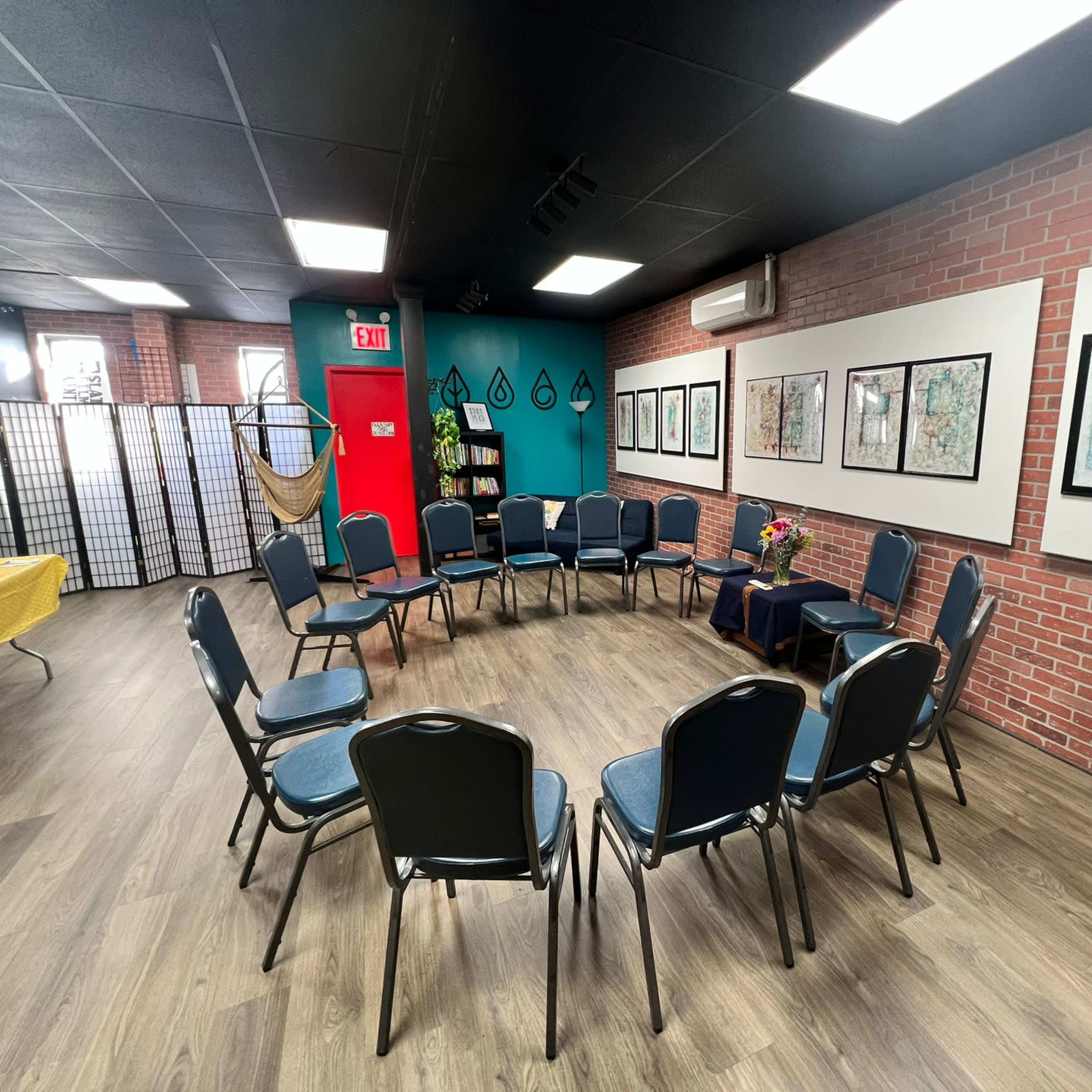 The room features a circular arrangement of blue chairs surrounded by framed artwork on the walls, with a partition and a table displaying a floral centerpiece in the background.