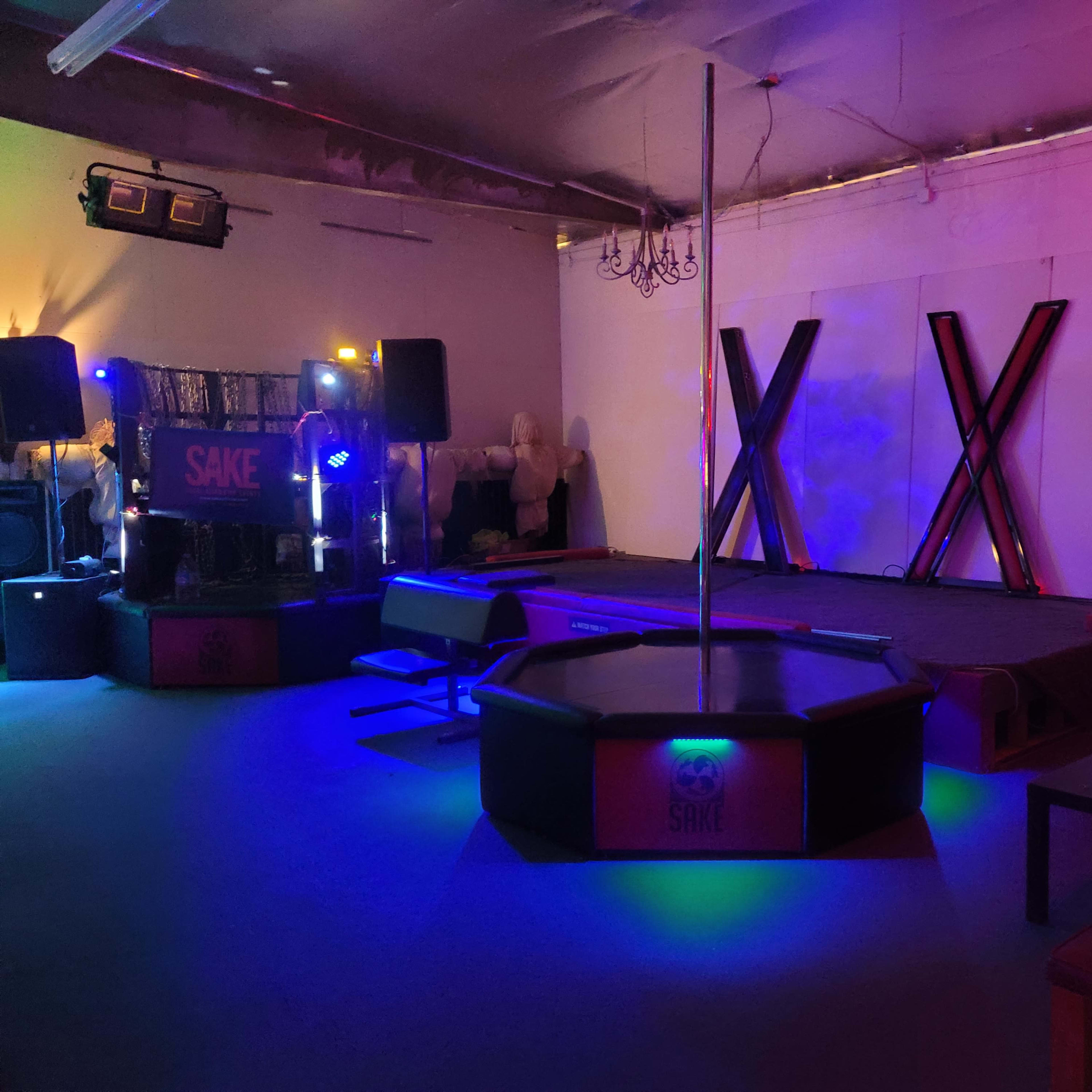 A dimly lit entertainment space with a stage featuring a pole, surrounded by seating and illuminated by colorful lighting.