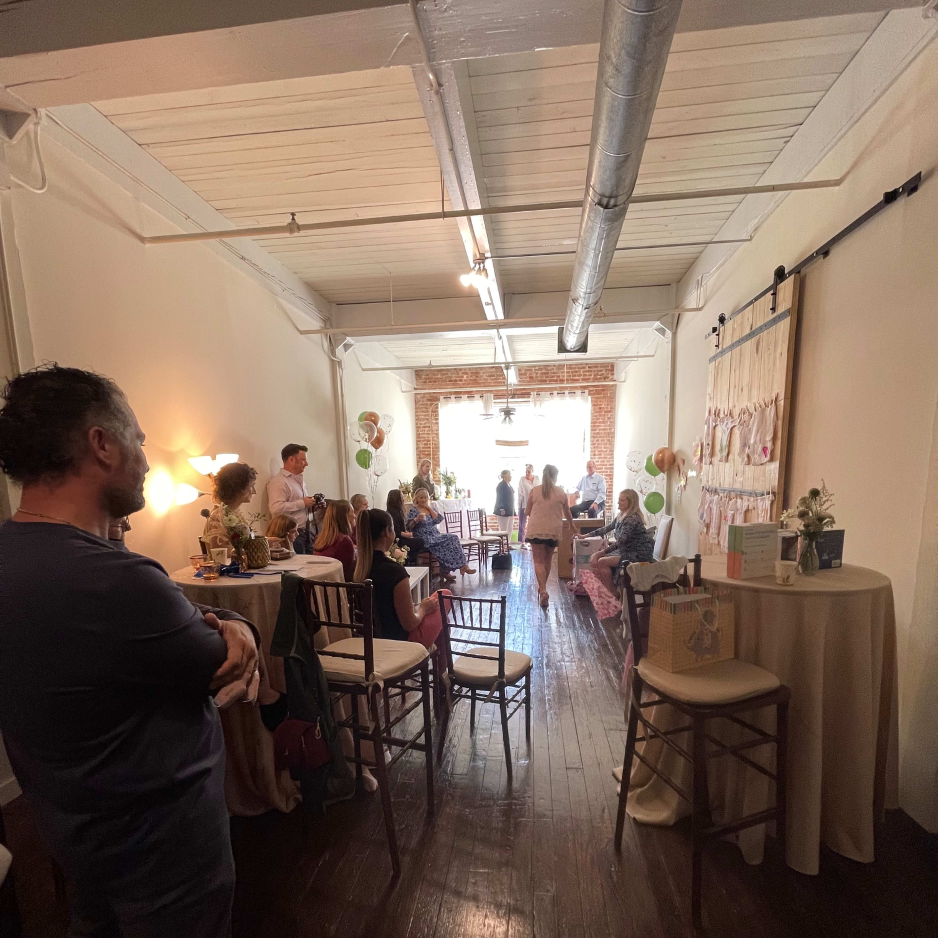 A group of people gathers in a spacious, warmly lit room with tables and decorations set up for an event.