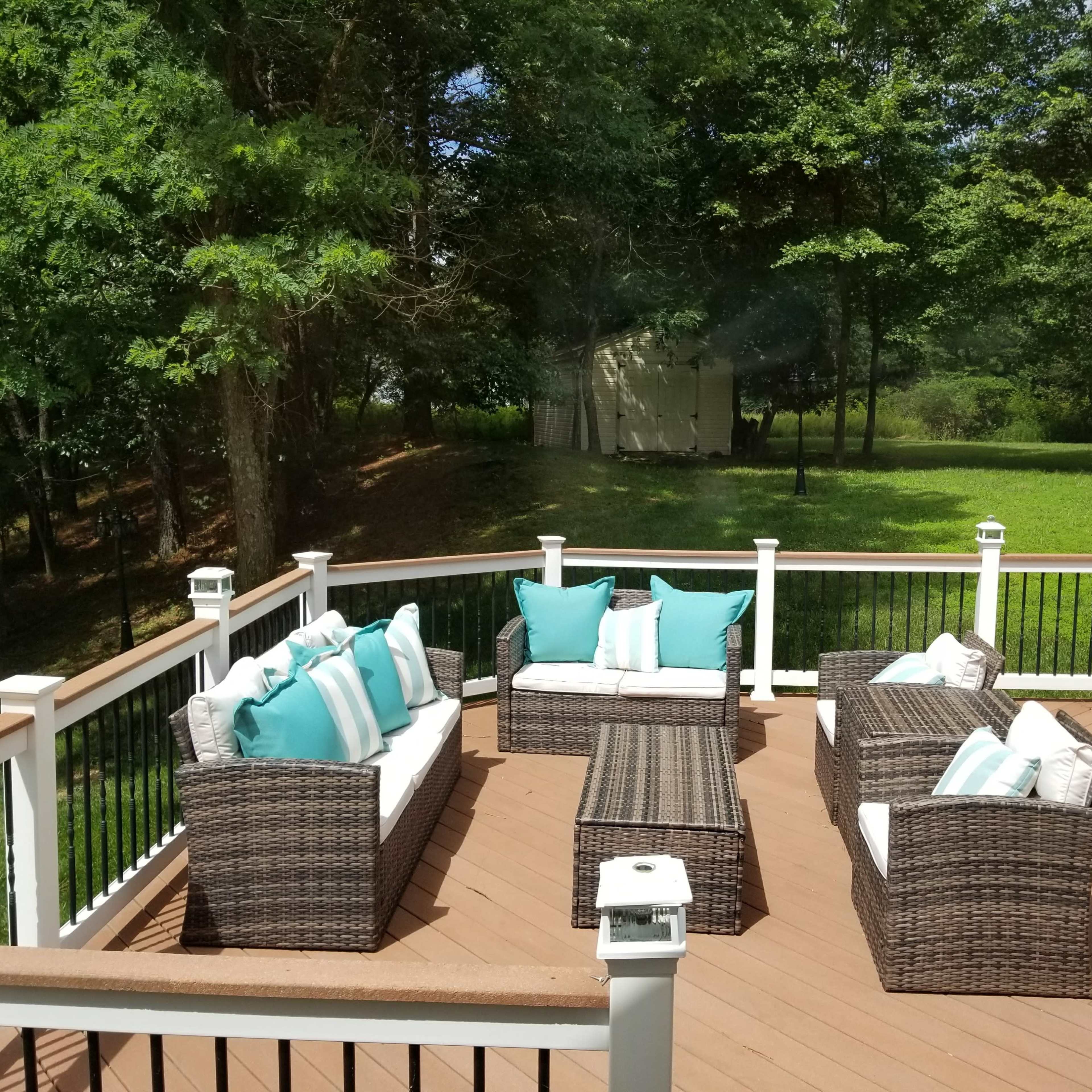 A deck features a seating area with wicker furniture adorned with turquoise and white cushions, surrounded by green trees and grass.
