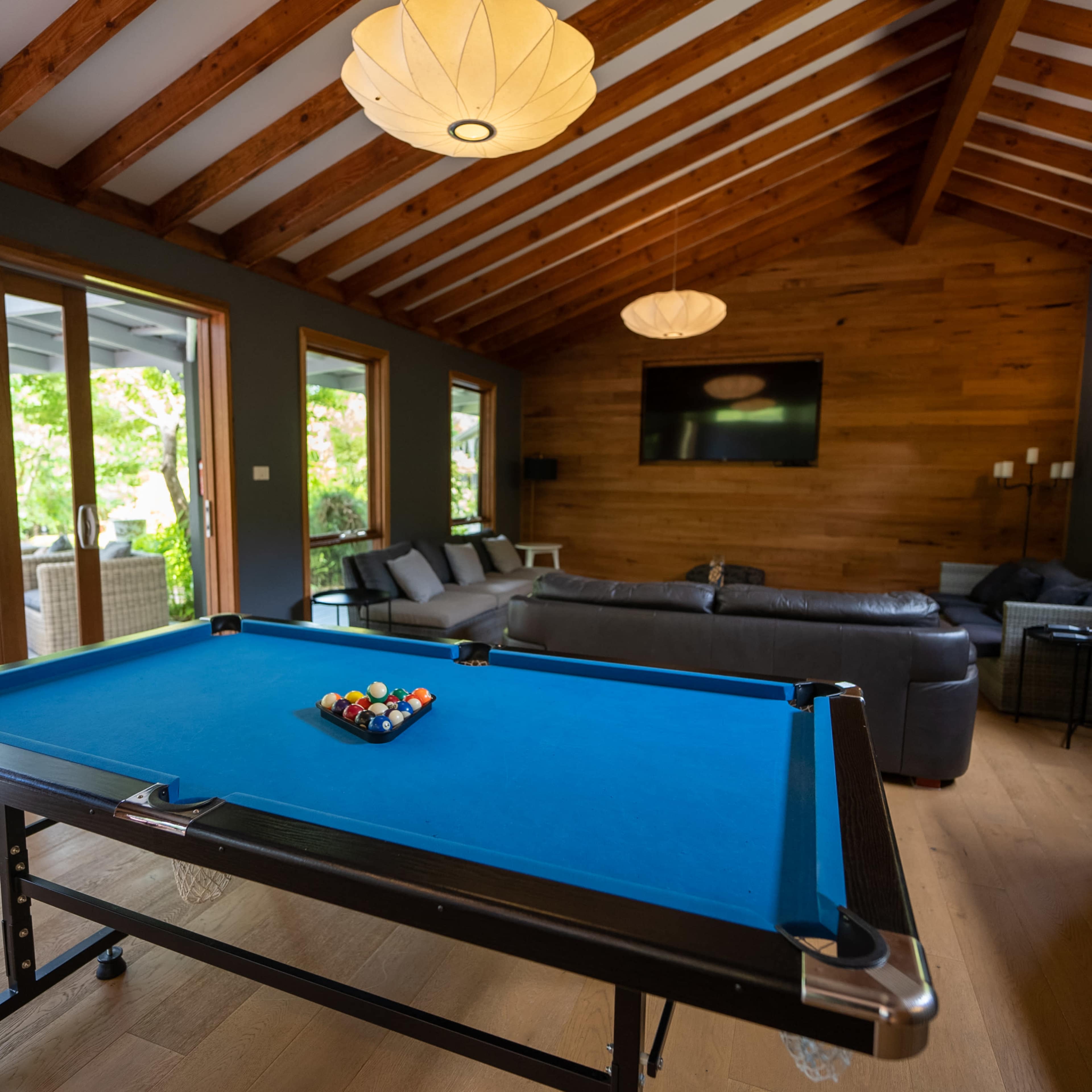 A blue pool table is positioned in a spacious room with wooden beams, surrounded by large windows and a seating area.