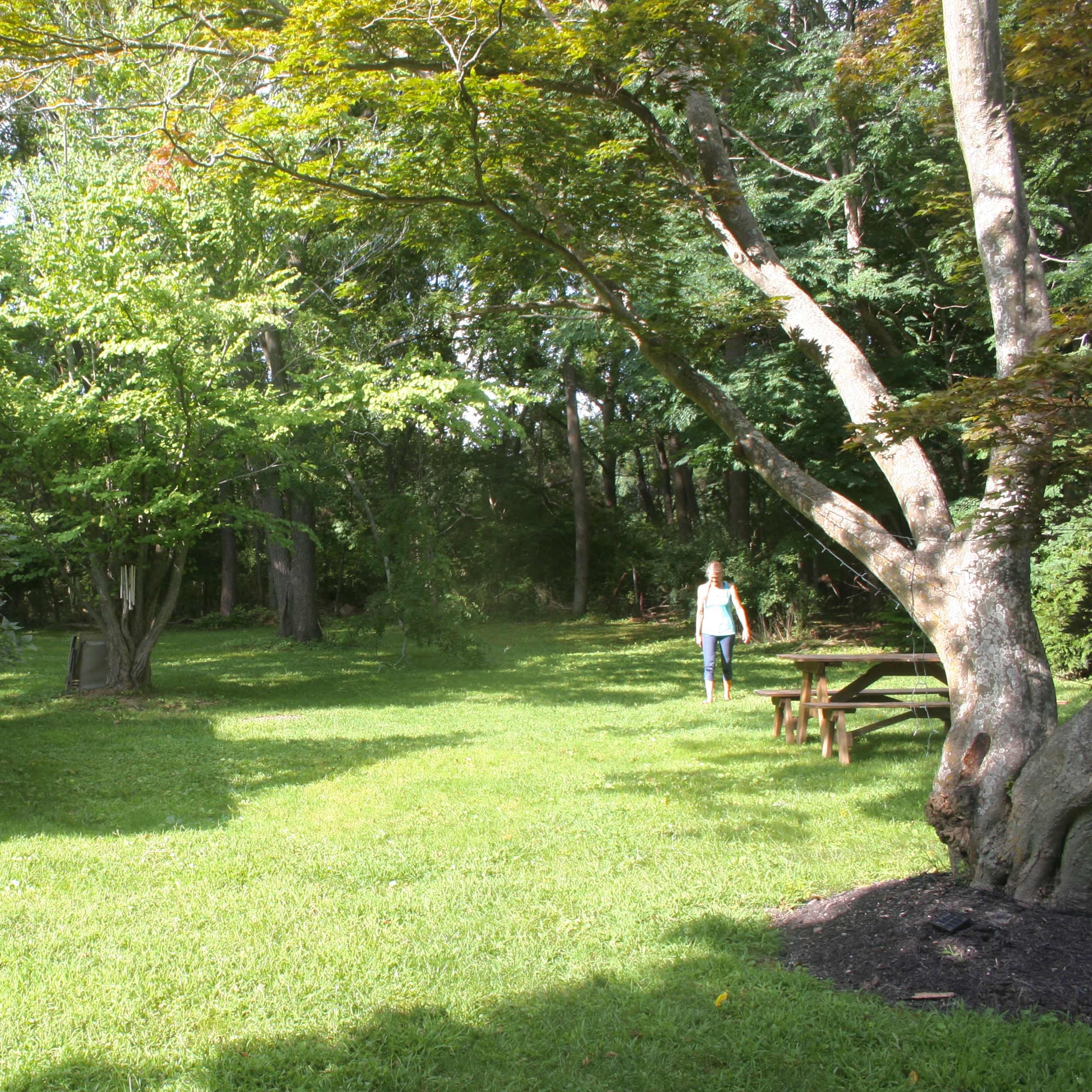 A woman walks through a grassy area near a small building and a picnic table surrounded by trees.