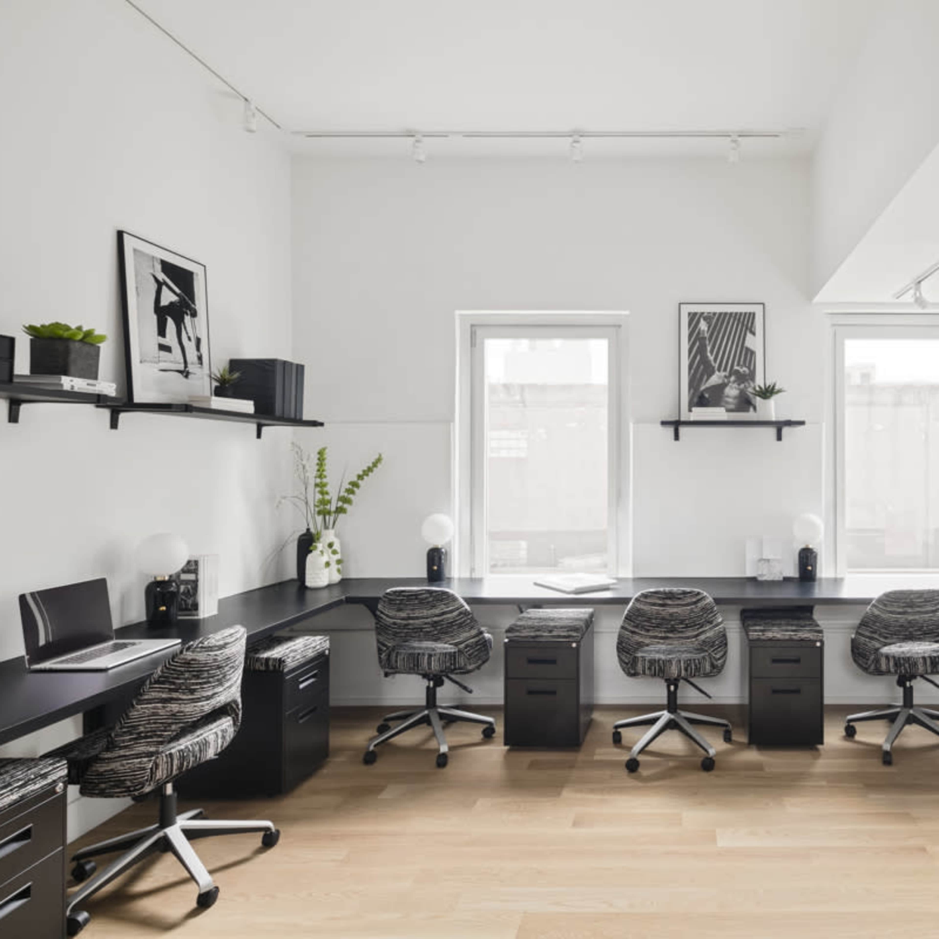 A modern office space features four ergonomic chairs around a U-shaped black desk with shelves holding decorative plants and framed artwork.
