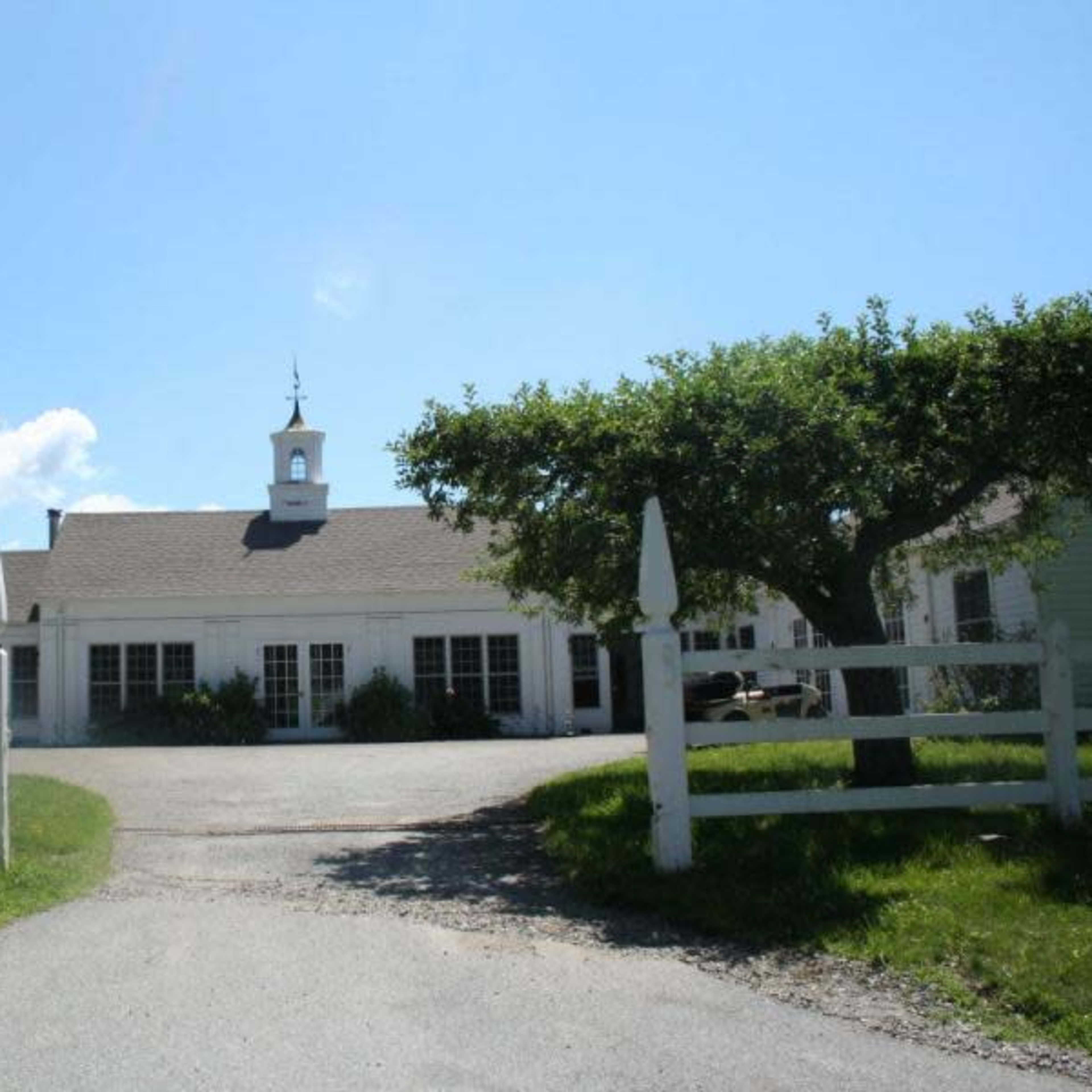 A white building with a steeple is surrounded by green trees and a gravel driveway, leading to a fenced entrance.
