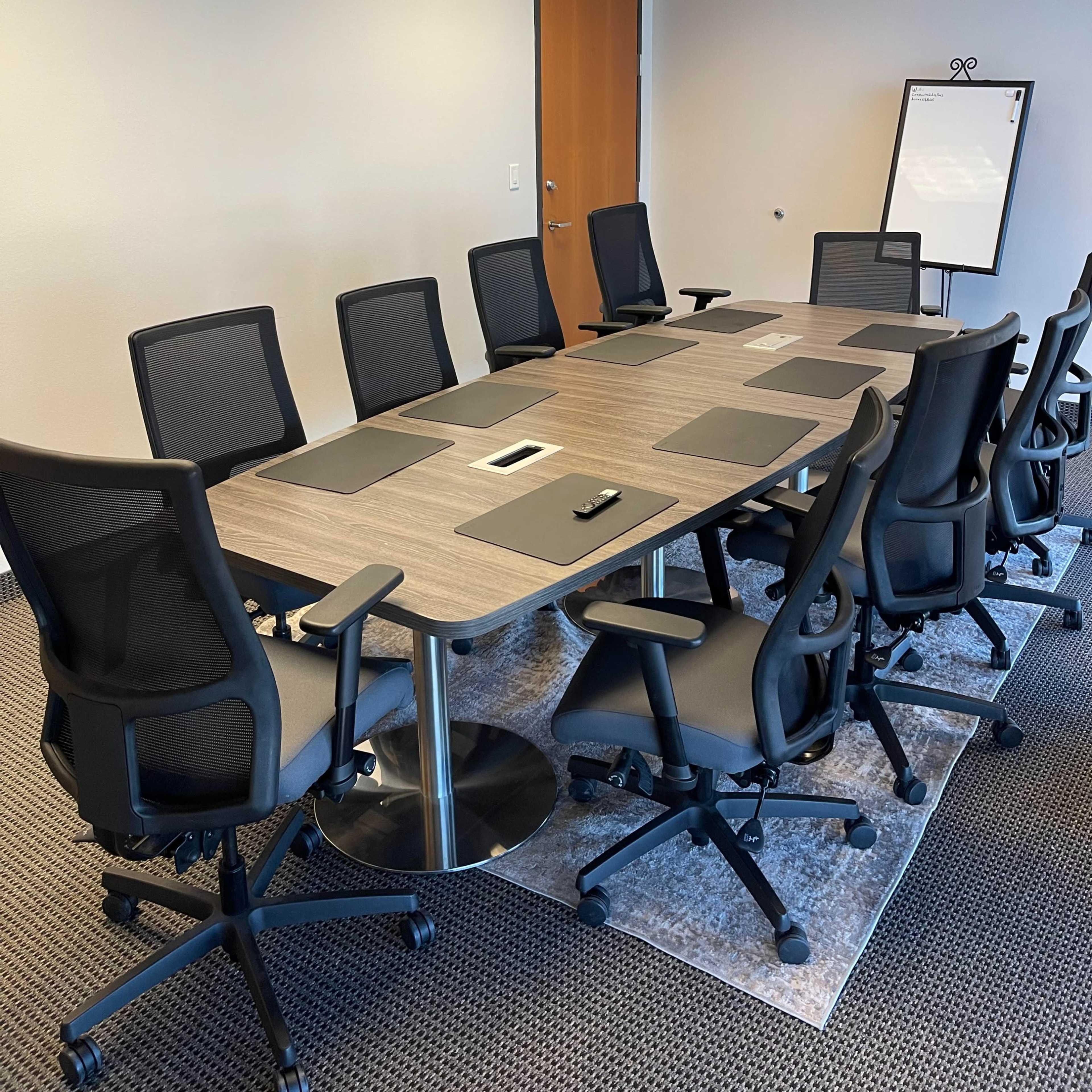 A modern conference room features a large table surrounded by ergonomic chairs, with a whiteboard and door visible in the background.