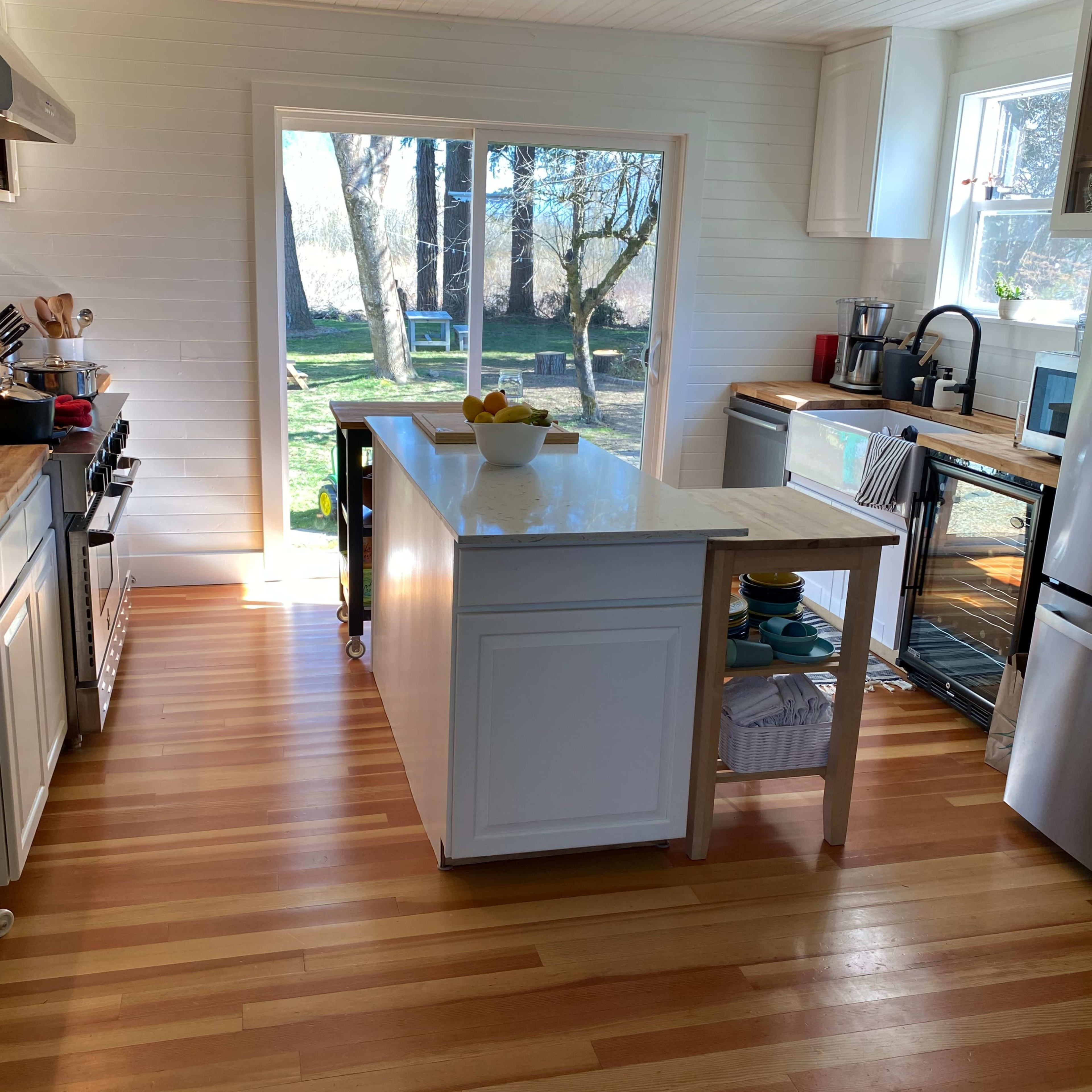 A bright kitchen features a central island, stainless steel appliances, and large windows overlooking a green outdoor area.