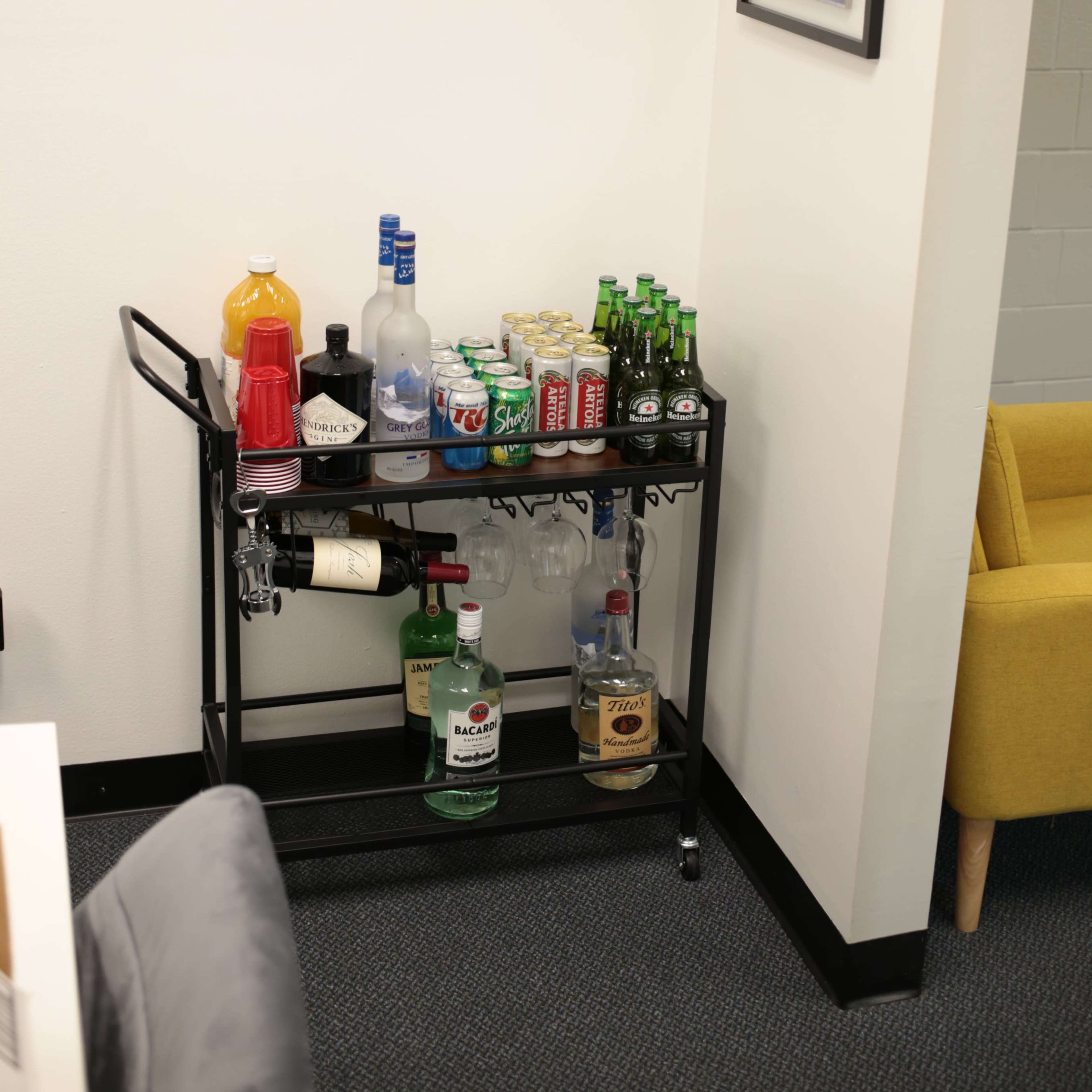A rolling bar cart holds a variety of alcoholic and non-alcoholic beverages against a wall in a room with light-colored walls and a framed poster.