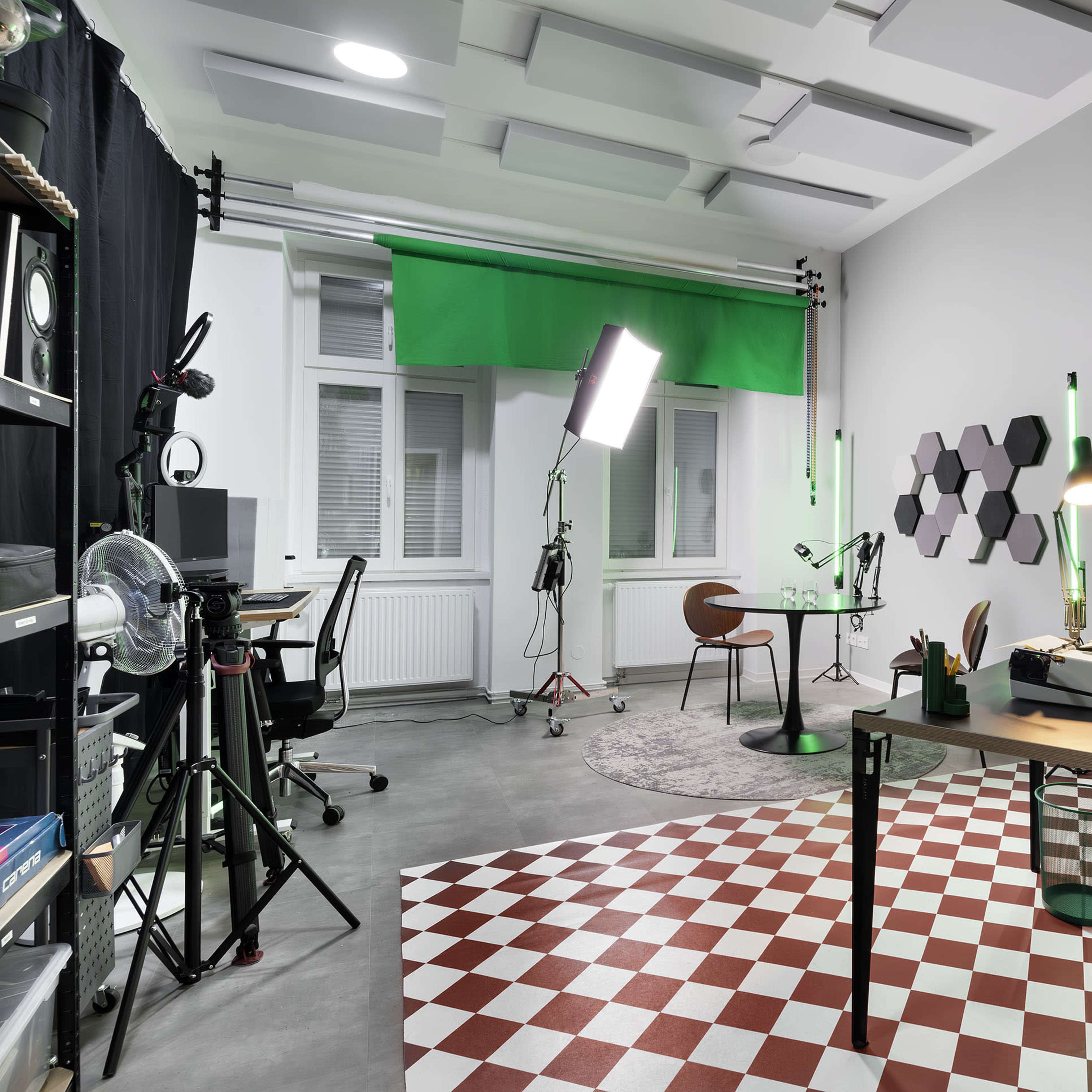 The scene depicts a modern studio workspace with a checkered floor, a desk with a typewriter, a green screen setup, and various potted plants.