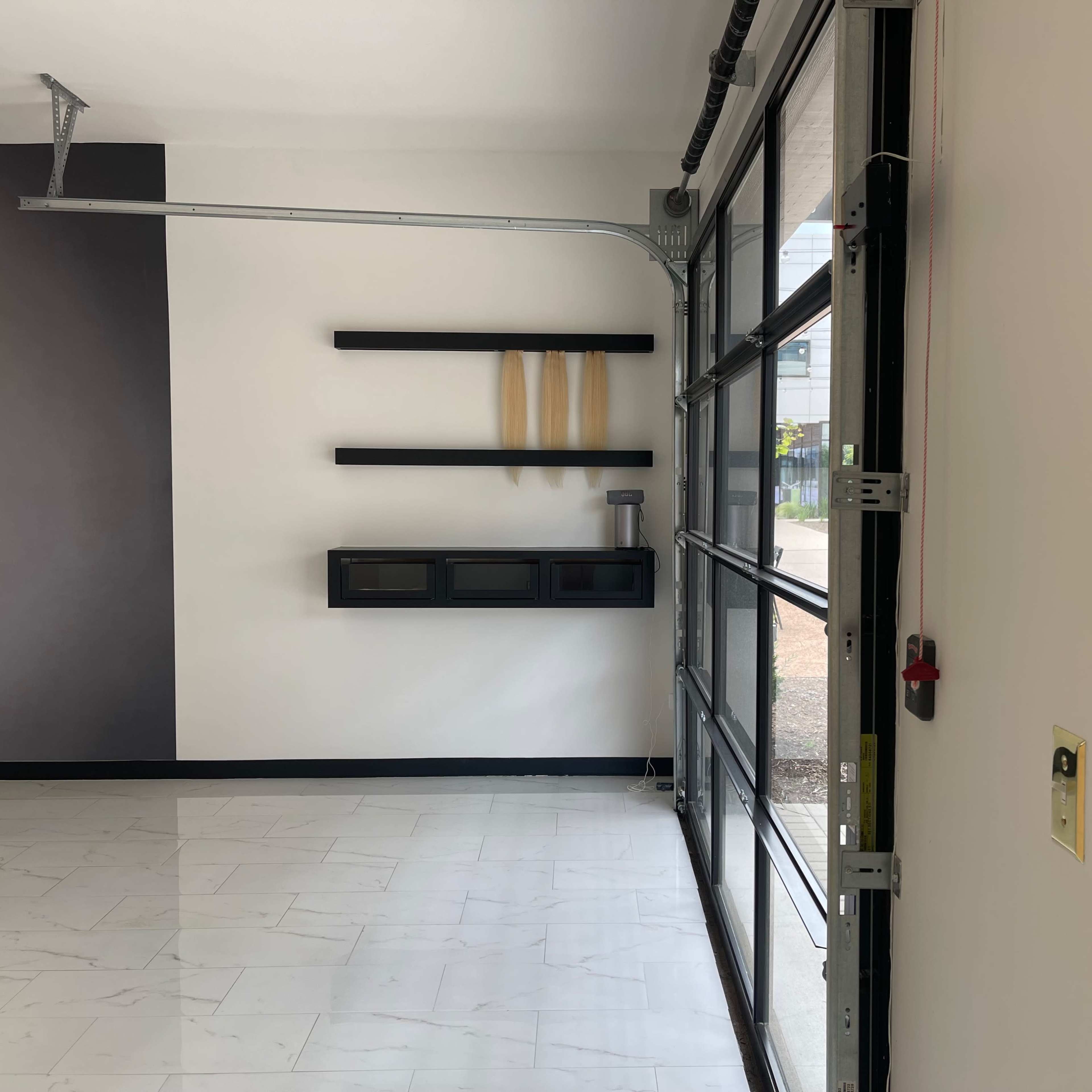 An empty room with a large glass garage door, white tiled flooring, and a black shelf mounted on the wall.