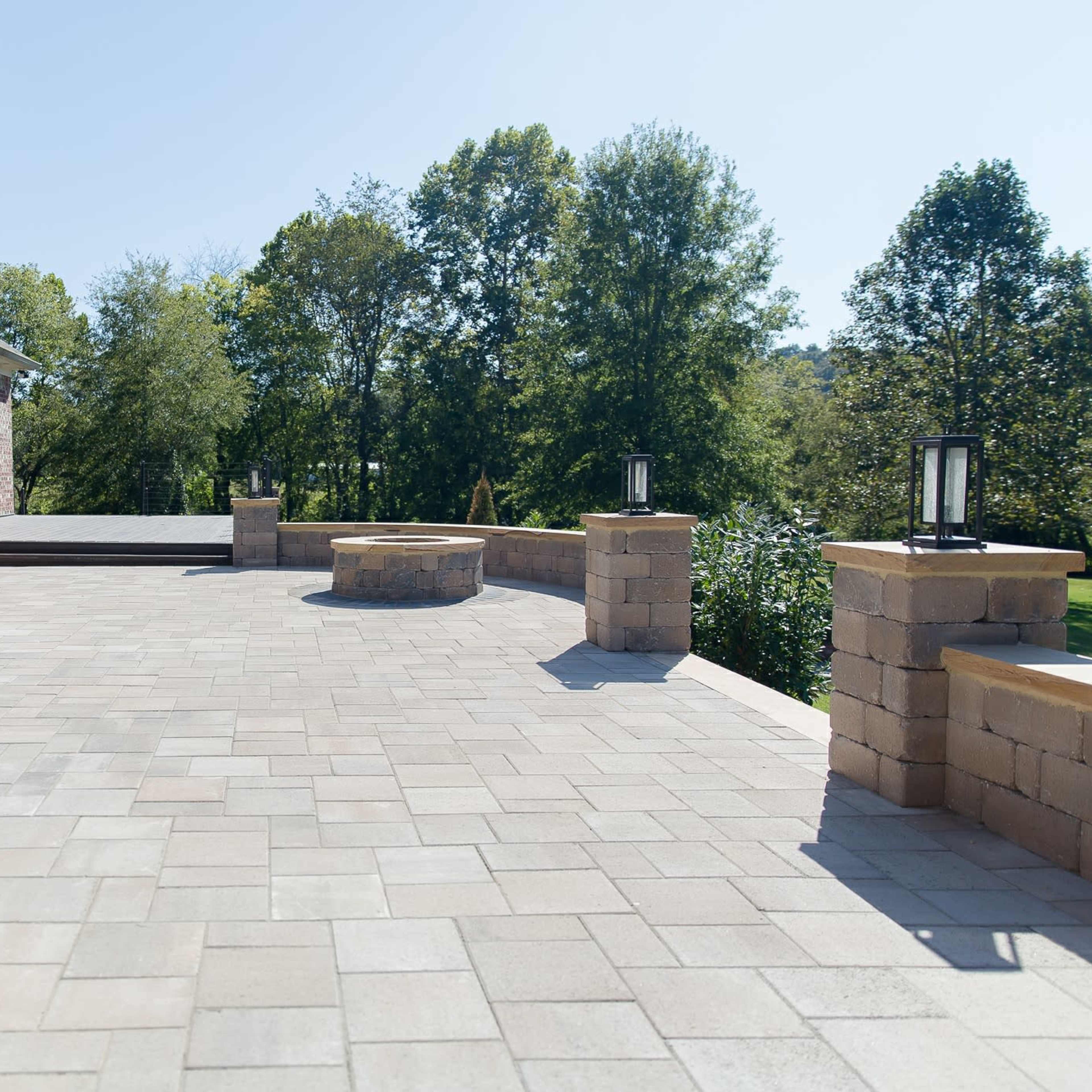 A spacious outdoor patio made of stone pavers, adjacent to a brick house, with a circular fire pit and surrounding greenery.