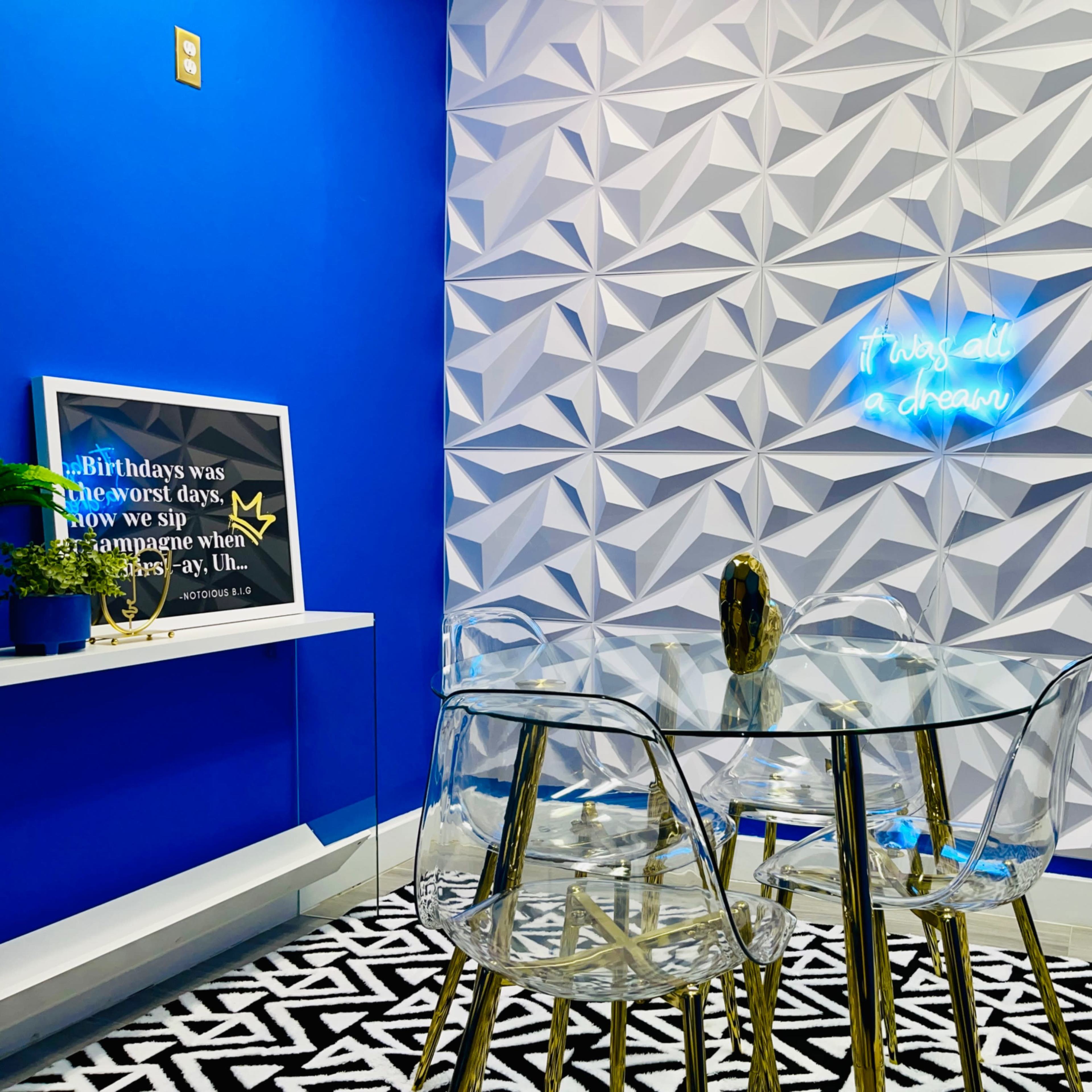 A glass table with transparent chairs is positioned against a textured blue and white wall featuring a neon sign that reads, "It was all a dream."
