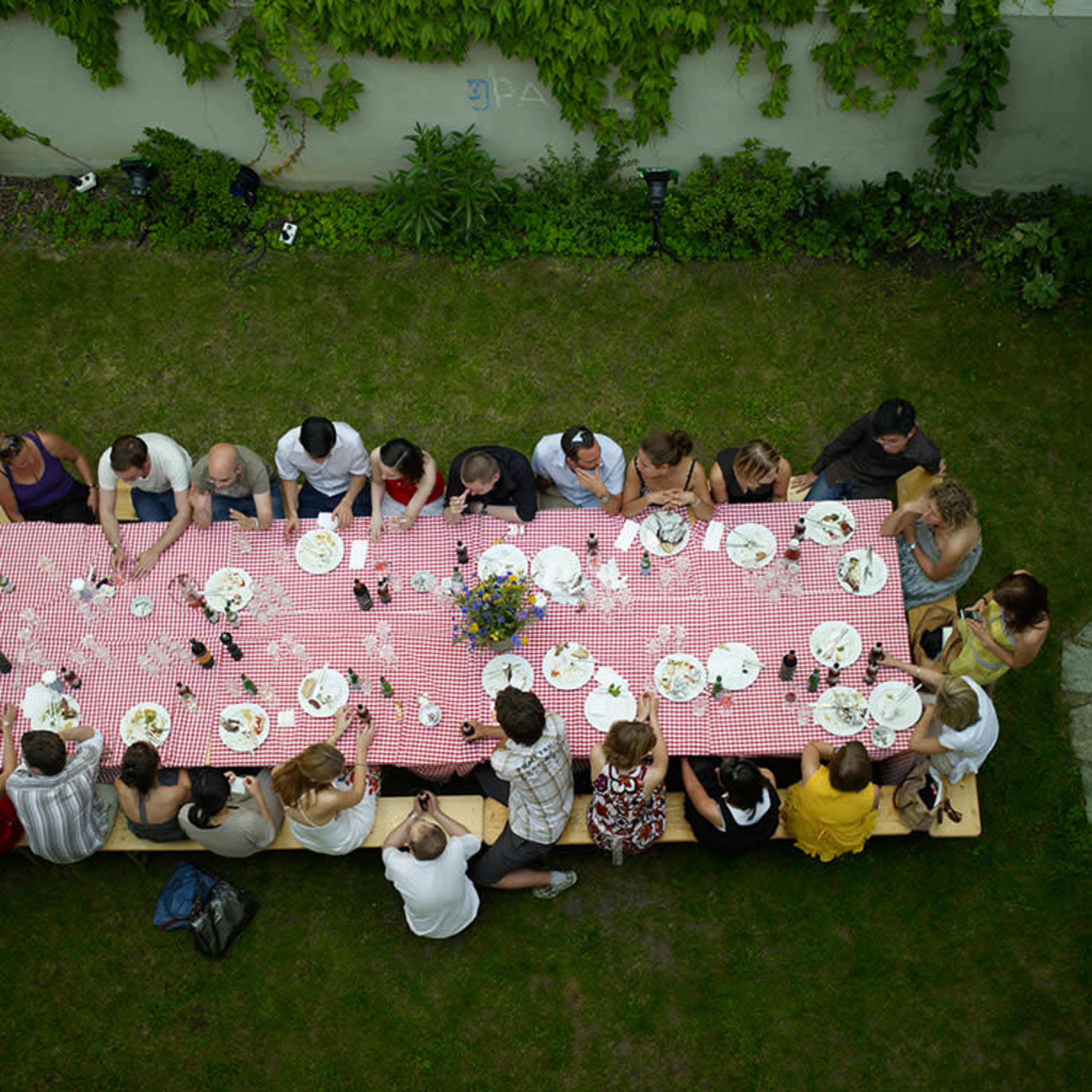 A long table covered with a red and white checkered tablecloth is filled with plates and drinks, surrounded by a group of people enjoying a meal outdoors.