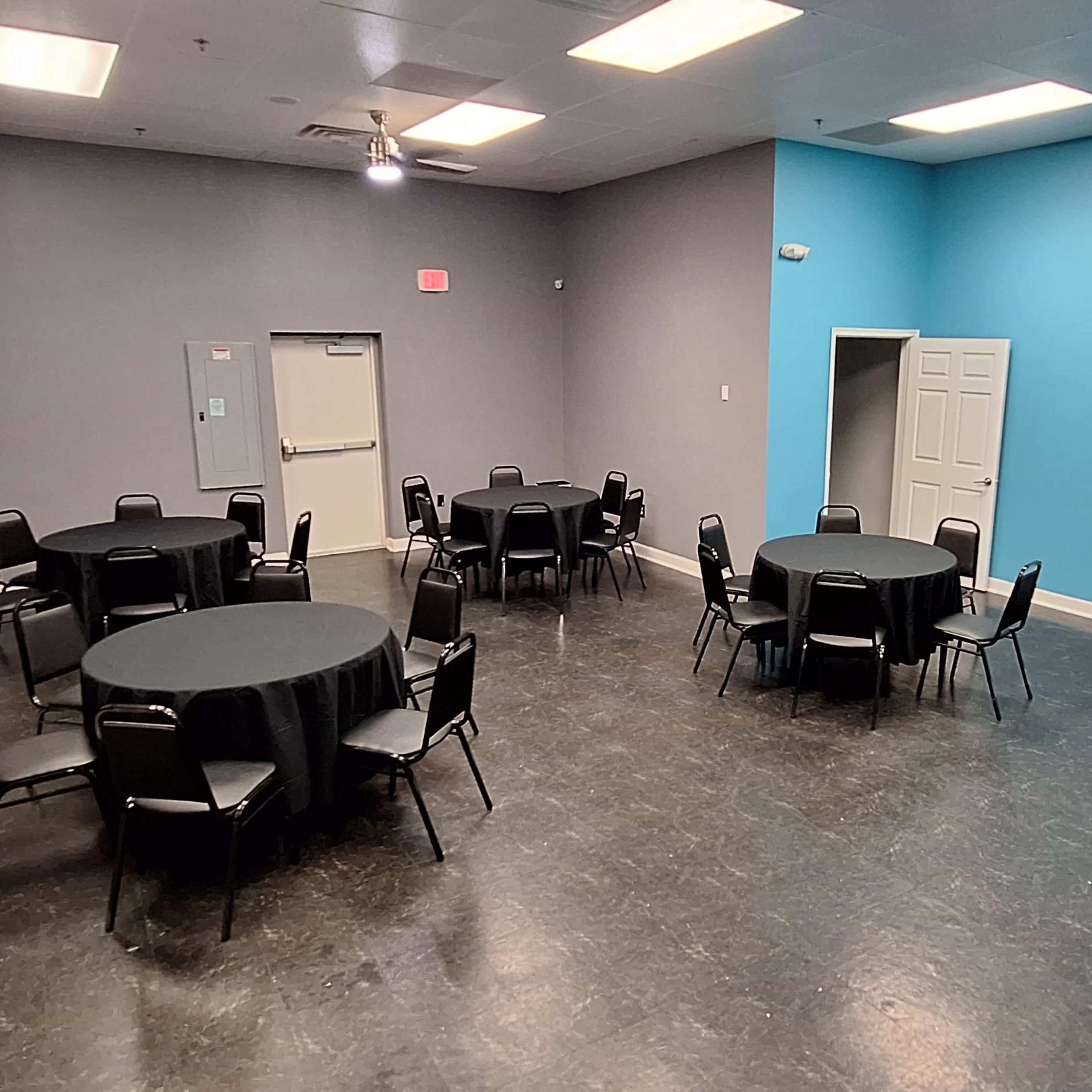 A spacious room with multiple round tables covered by black tablecloths, arranged around the space with a blue accent wall and grey surrounding walls.