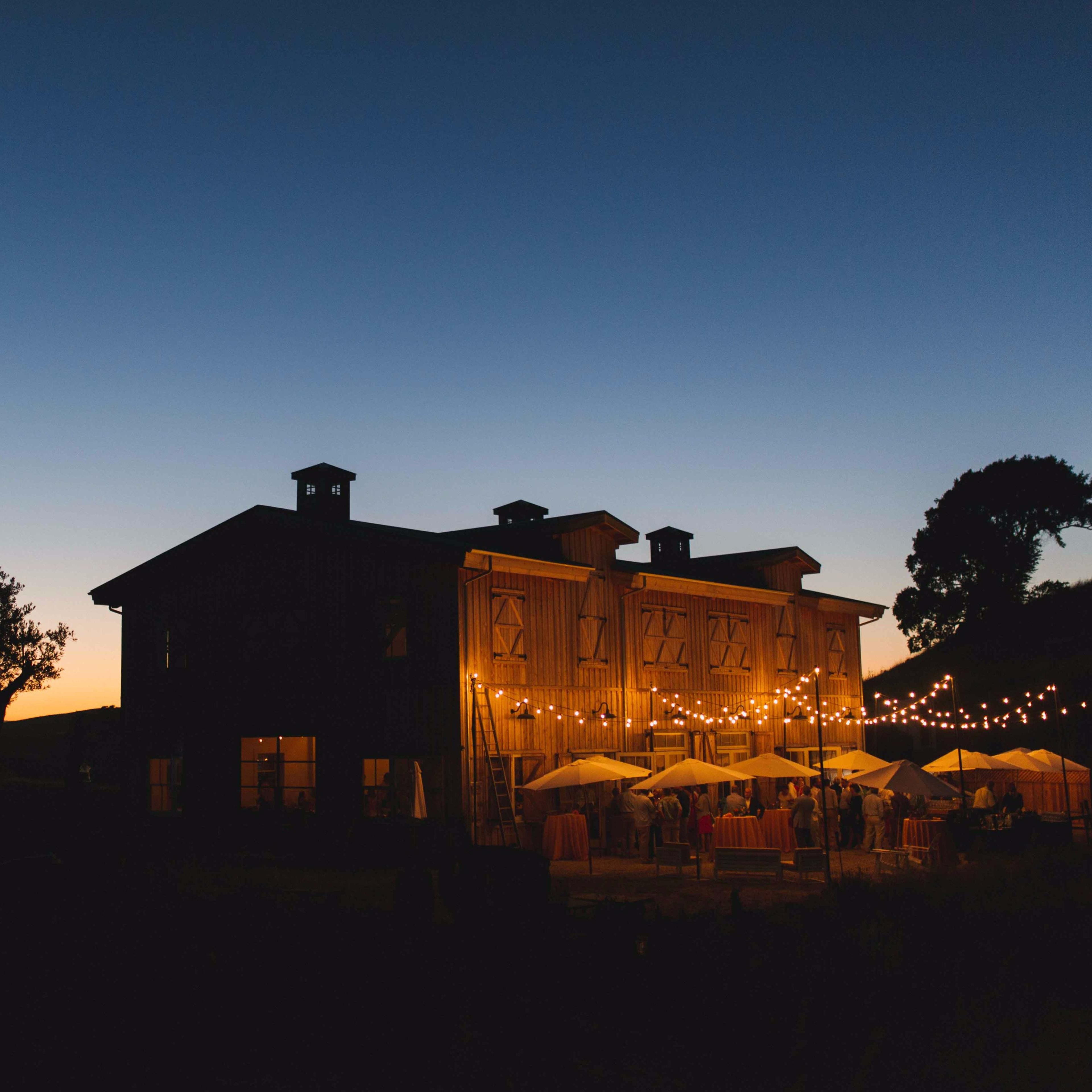 A rustic barn illuminated by string lights stands against a twilight sky with a sunset in the background.