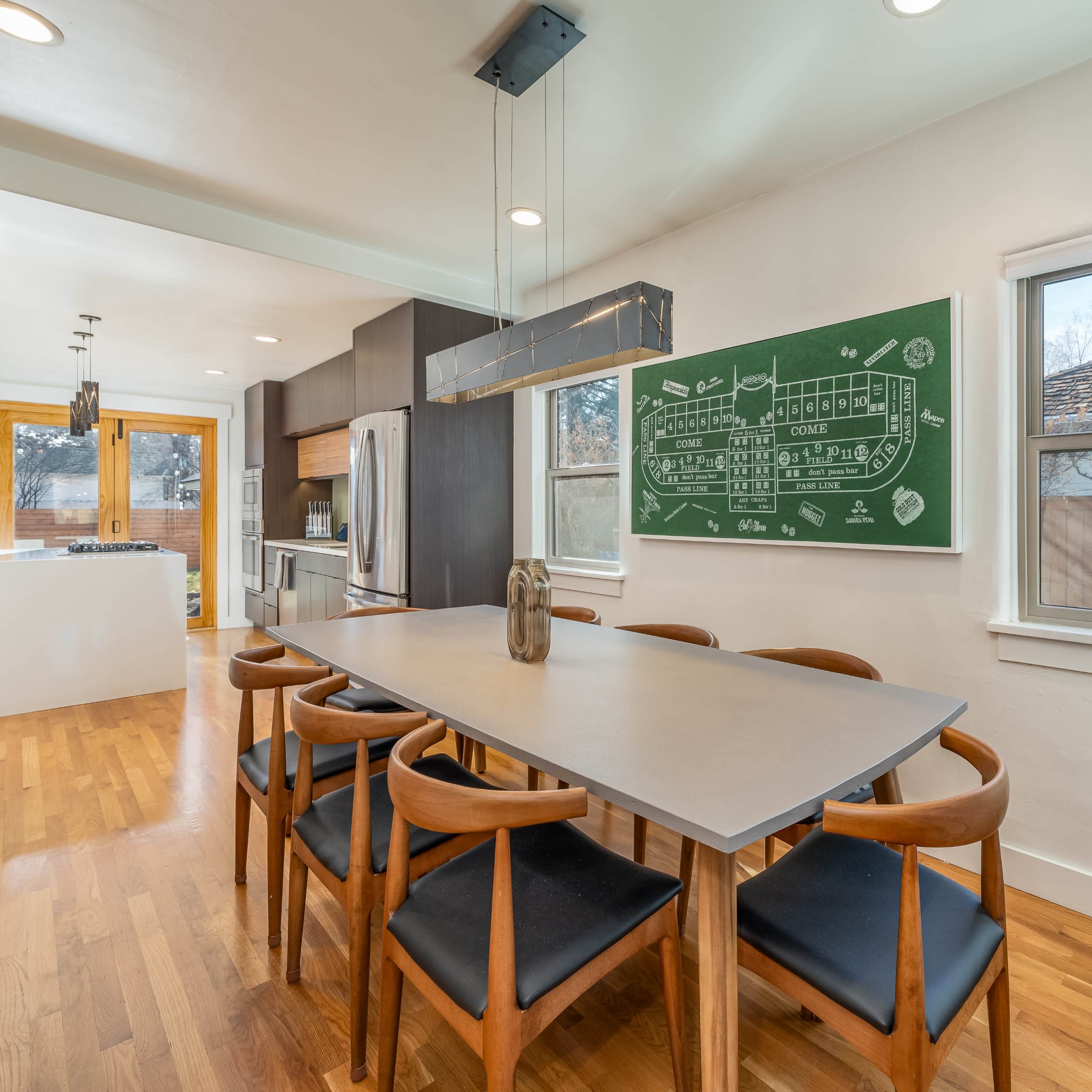 A modern dining area features a long table with wooden chairs, a chalkboard wall with a diagram, and large windows letting in natural light.