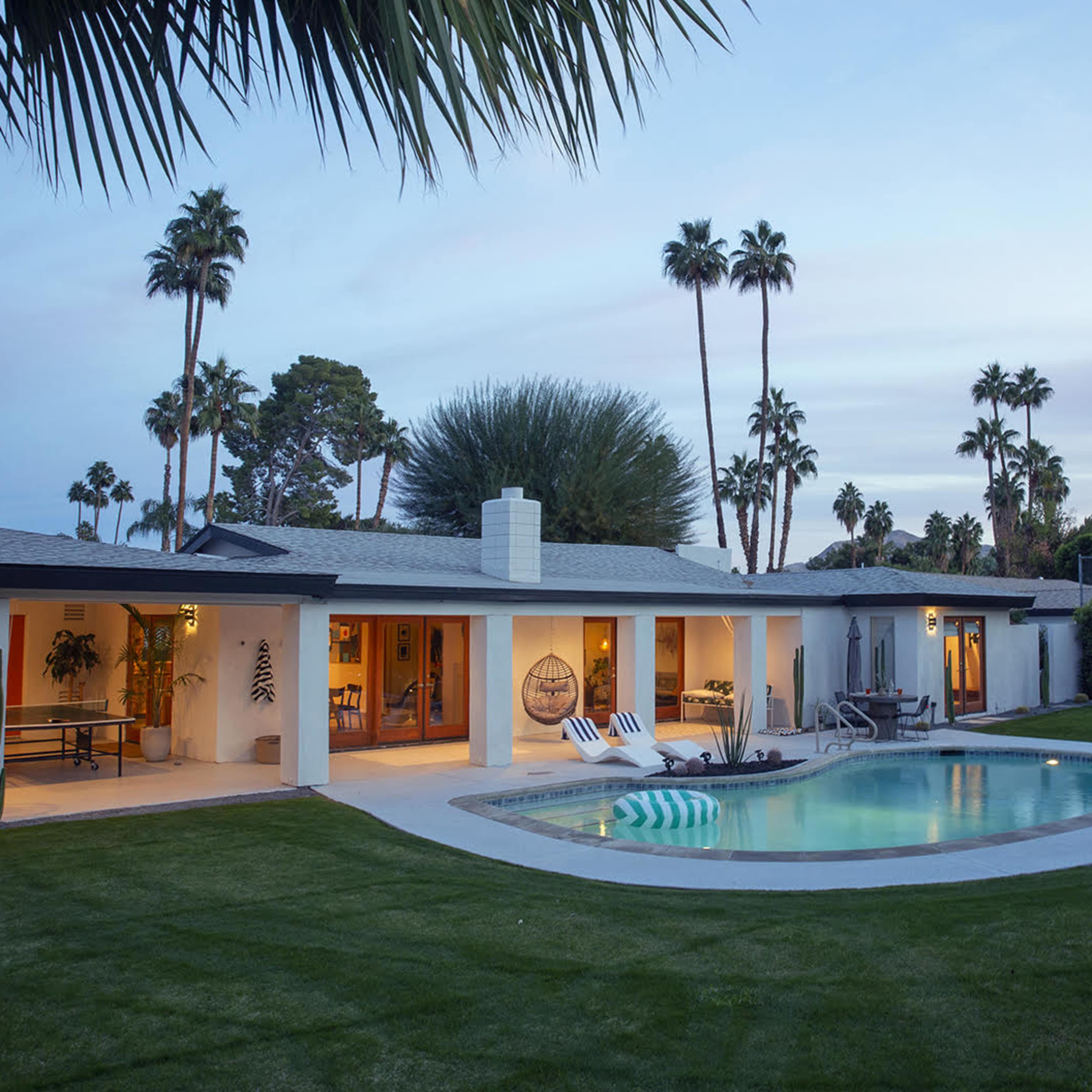 A modern house with a pool is surrounded by palm trees and lush green grass during twilight.