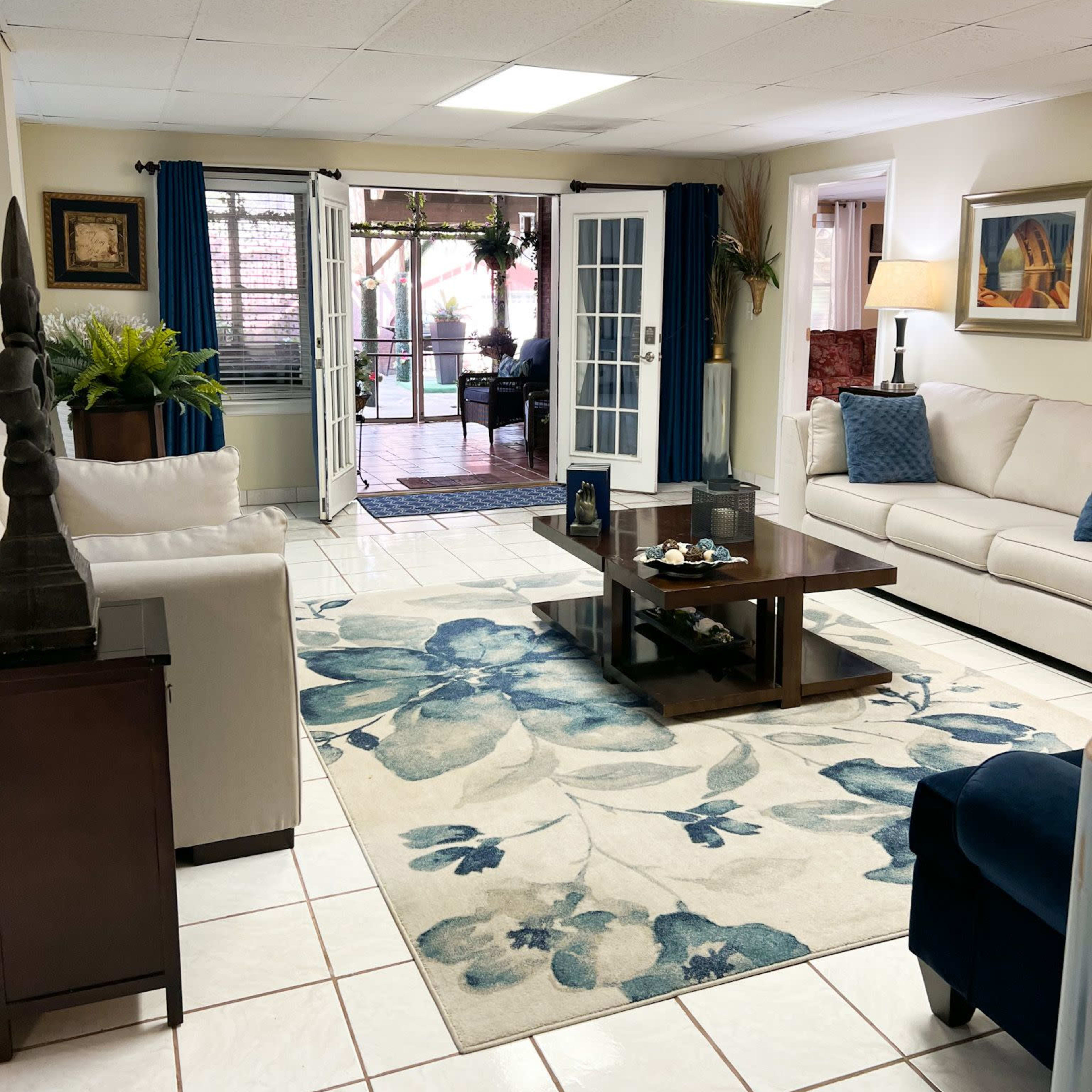A spacious living room with a white couch, a wooden coffee table, and a blue floral area rug, leading to a patio area visible through double French doors.