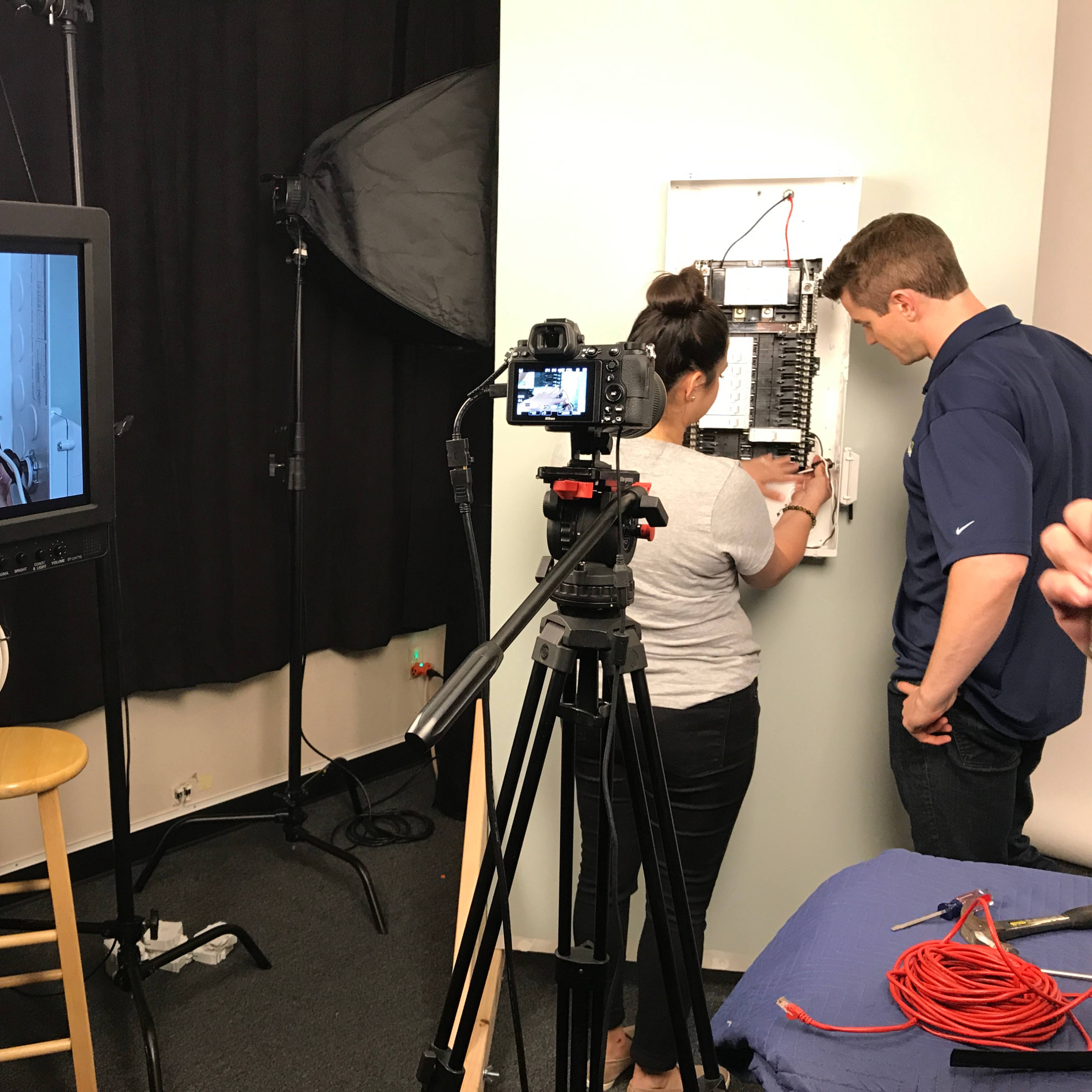A person works on an electrical panel while another films the process with a camera.