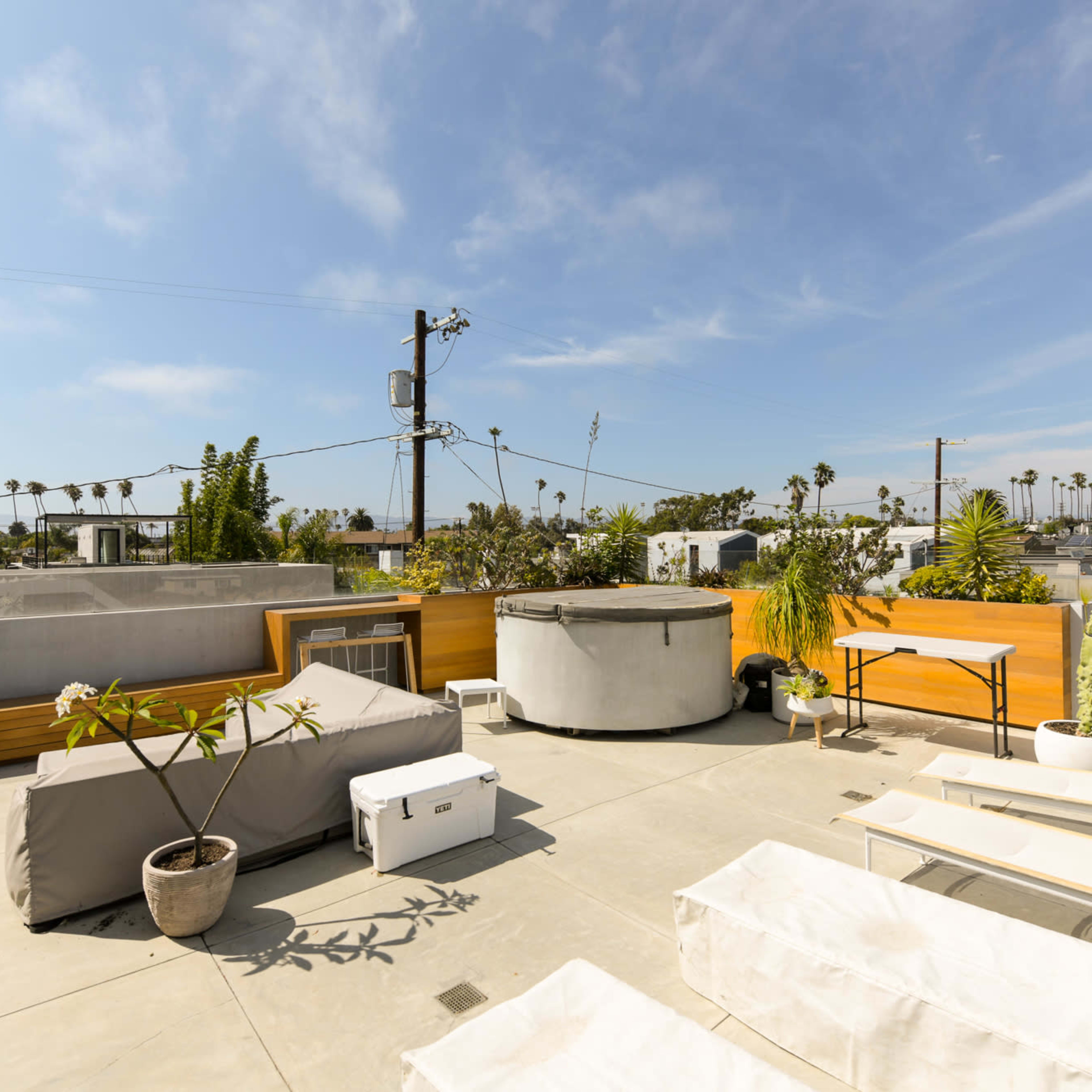 The rooftop terrace features wooden seating, a round hot tub, and several potted plants against a backdrop of palm trees and blue sky.