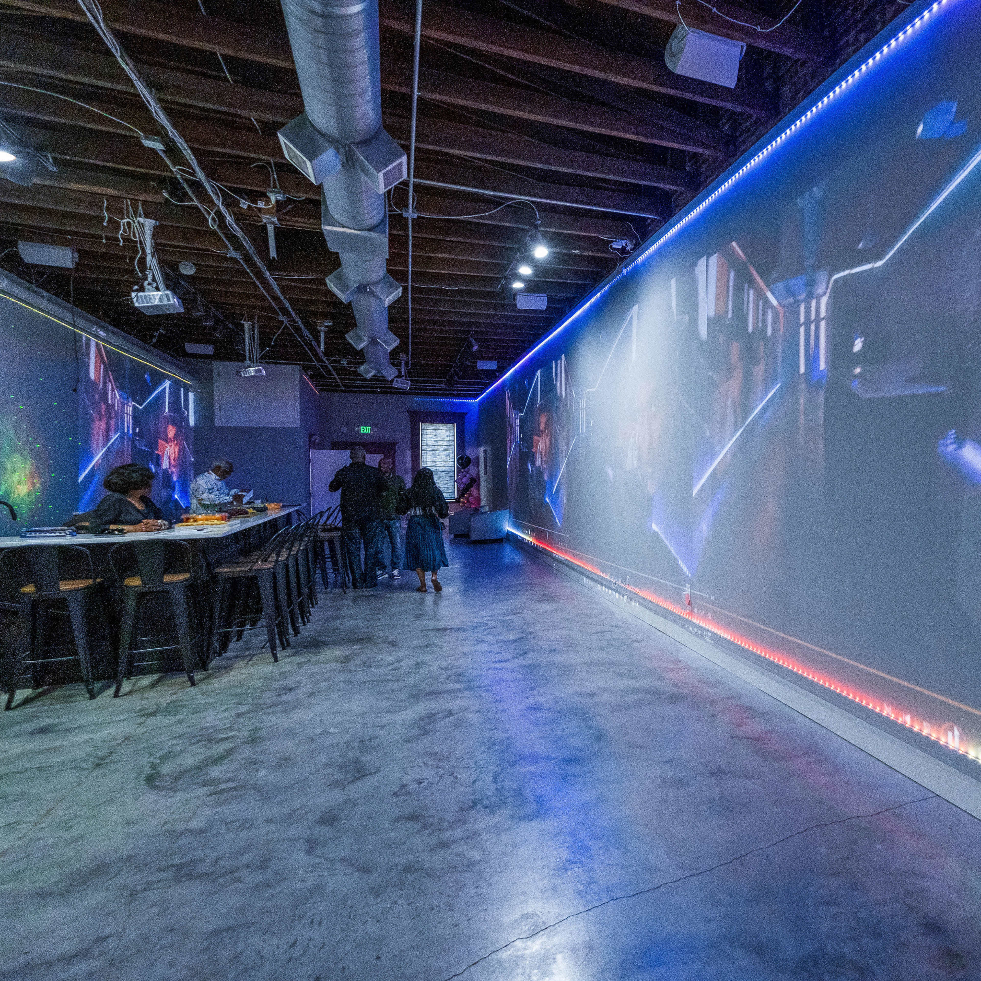A spacious indoor area with a long wall displaying vibrant digital screens and several people seated at high tables.