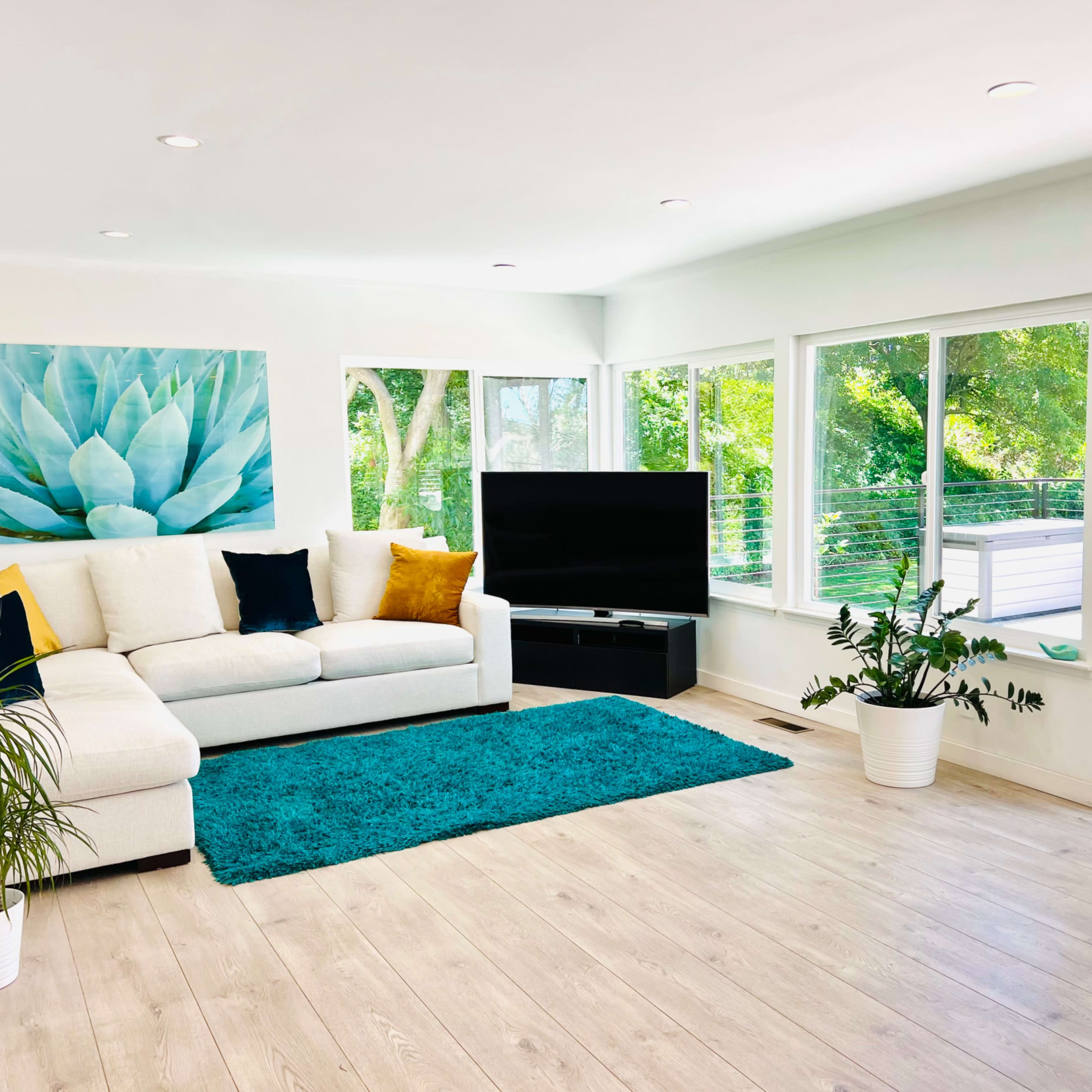 The living room features a white sectional sofa, a large black television, and a vibrant blue area rug on light wooden flooring, with large windows allowing natural light and views of greenery outside.