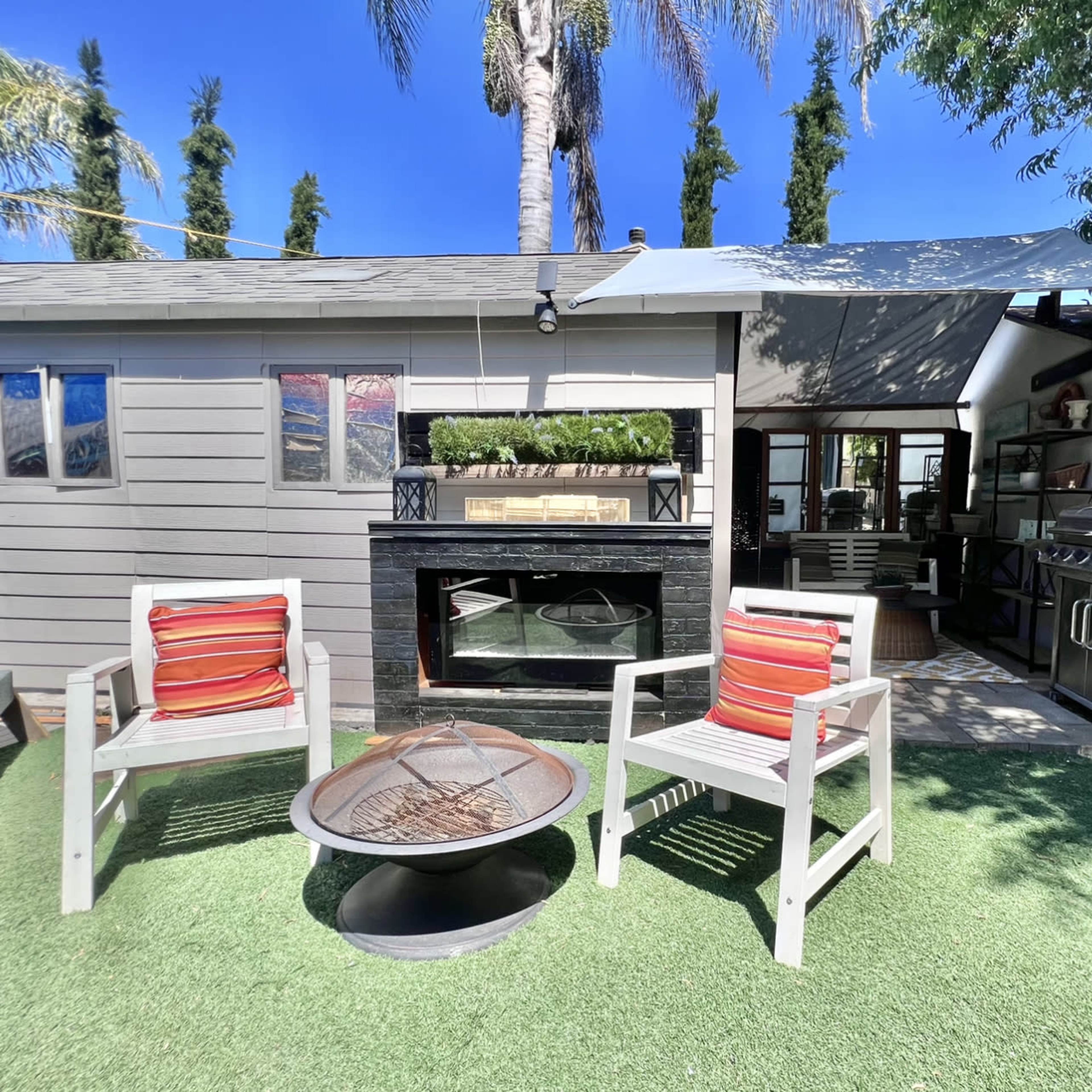 A small outdoor patio features two striped chairs, a fire pit, and a well-equipped kitchen area adjacent to a gray-shingled house.