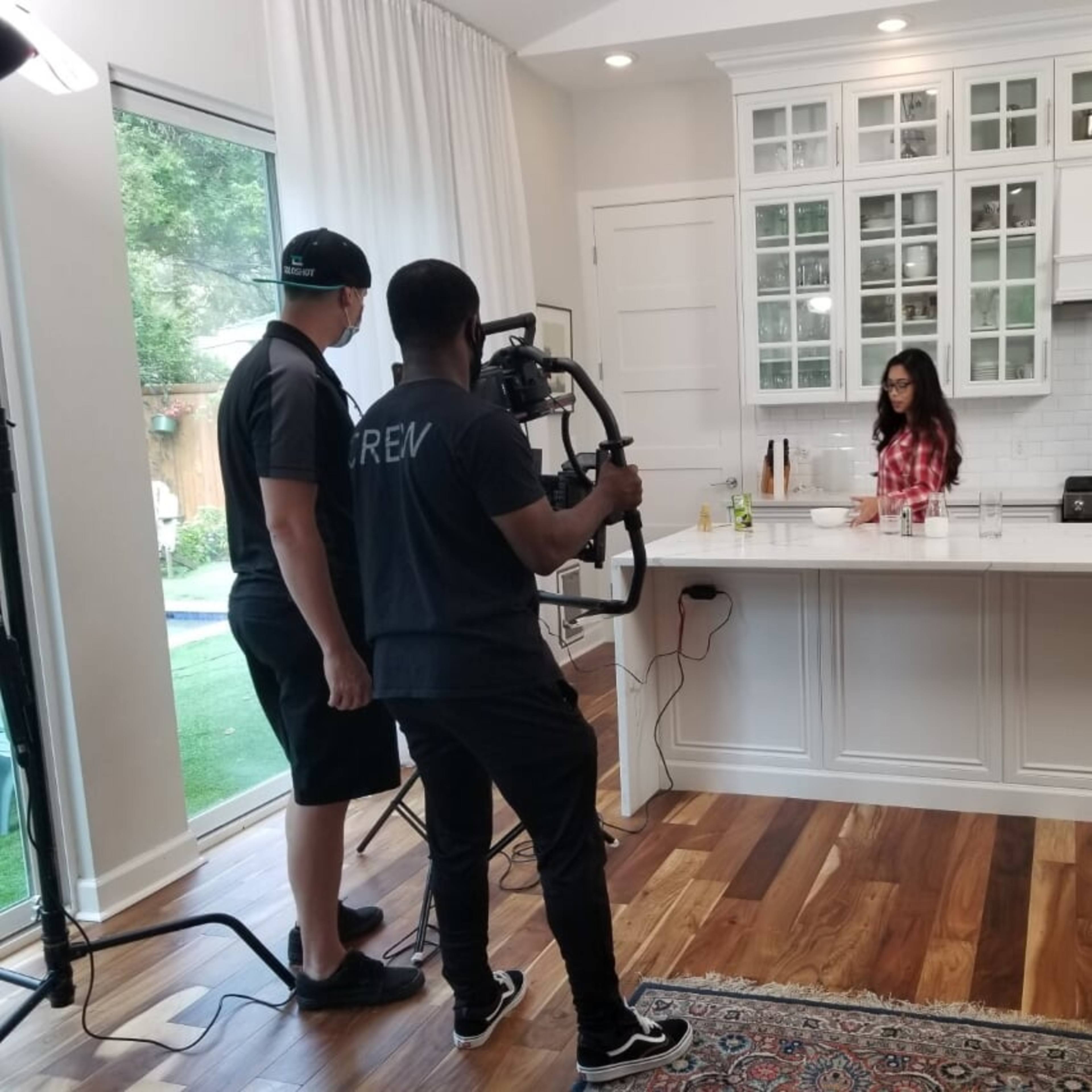 A woman prepares food at a kitchen island while two crew members film the scene with a camera setup.
