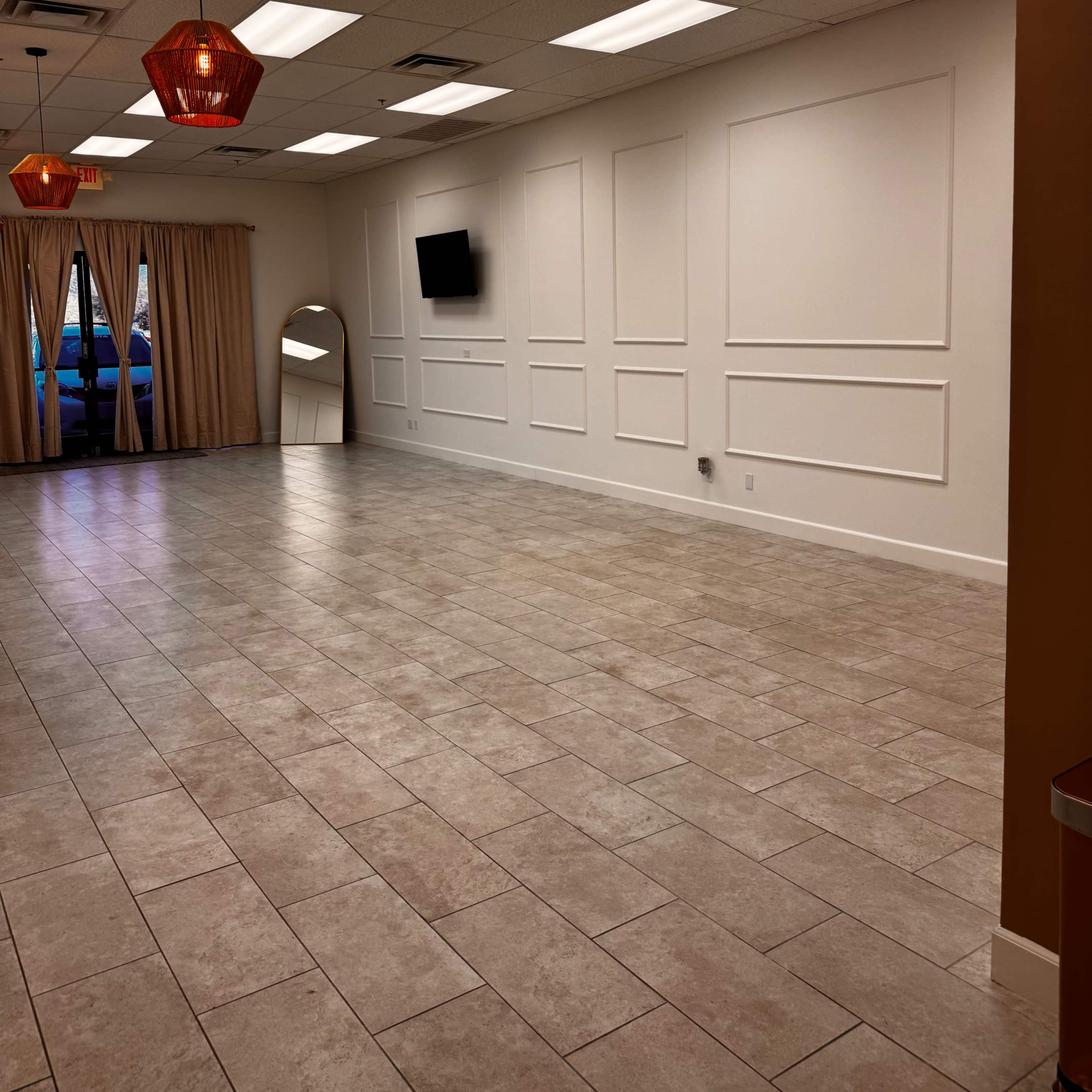 A spacious, well-lit interior with tiled flooring, white walls, and decorative lighting fixtures, featuring a large mirror and a television mounted on the wall.
