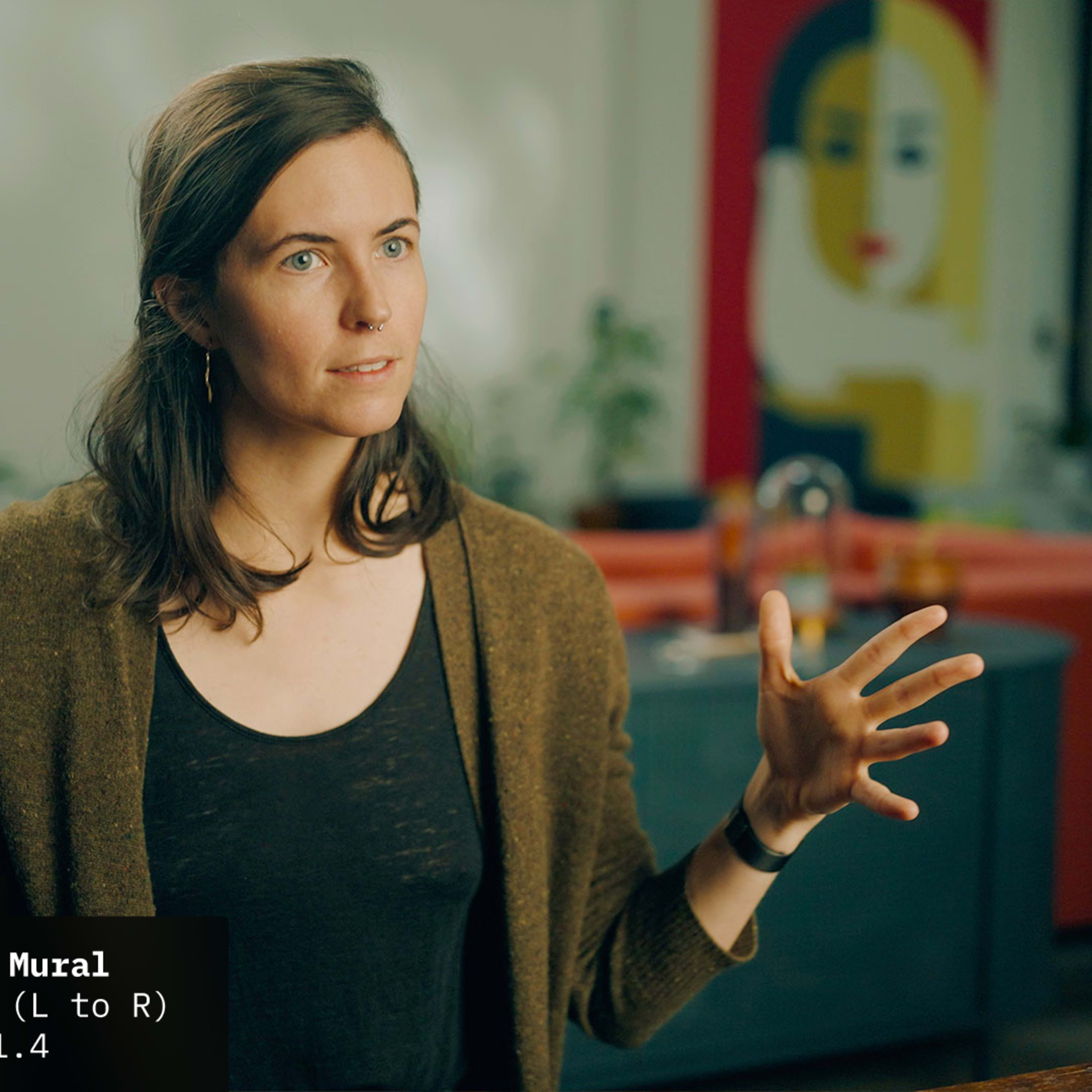 A woman is gesturing with her hands while speaking, seated at a table in front of a colorful mural.