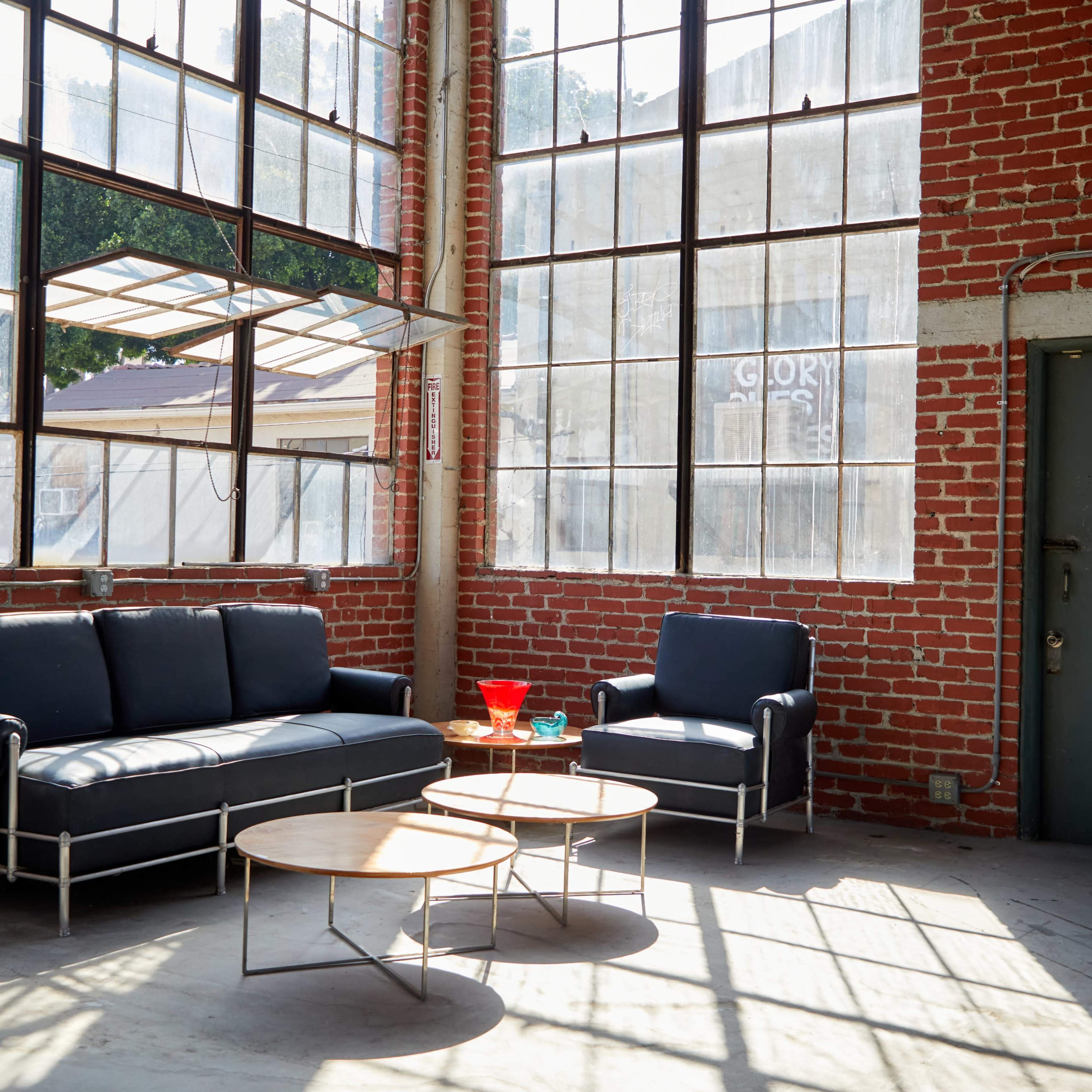 A modern seating area with a black leather sofa and two round wooden coffee tables, situated in a space with brick walls and large windows.