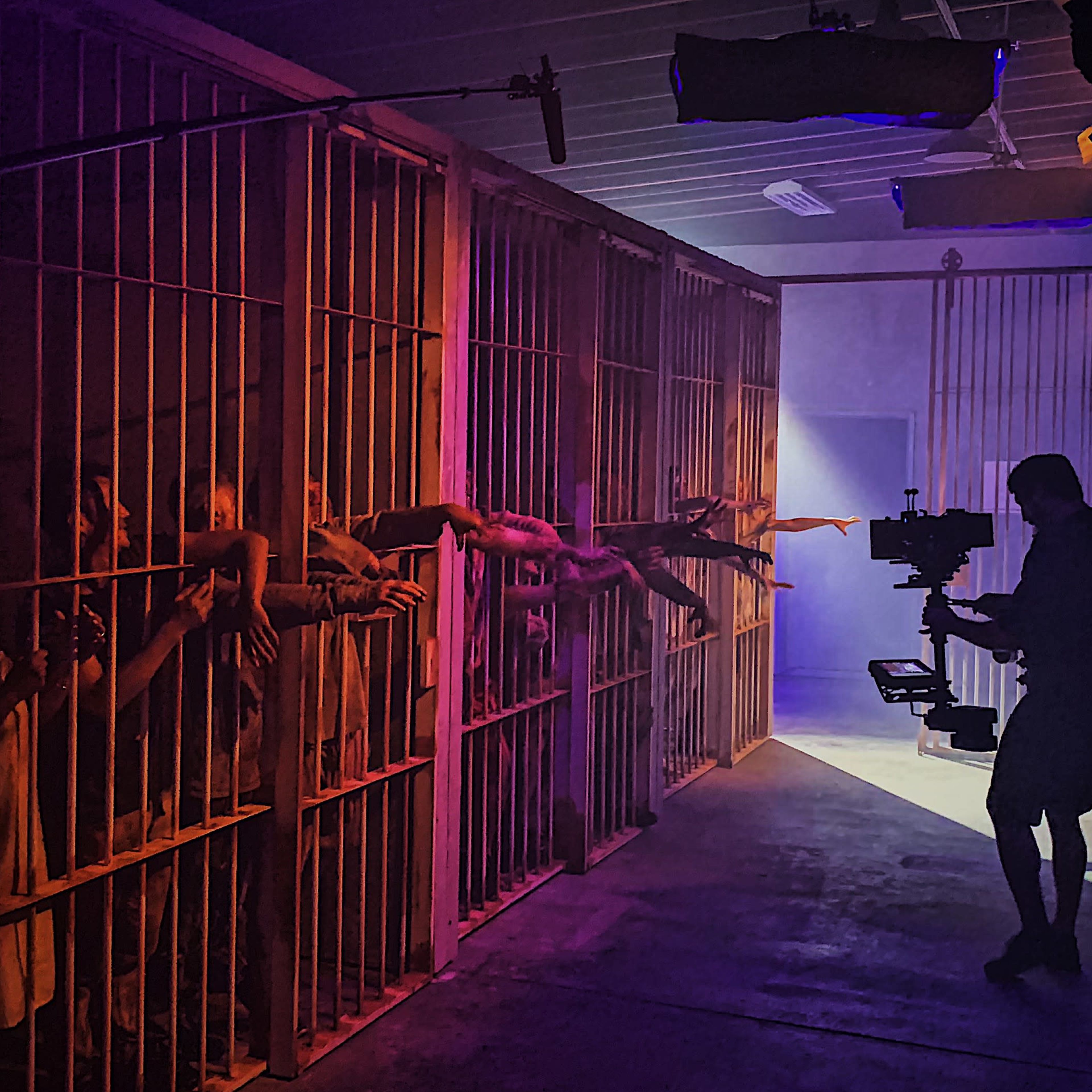 A cinematographer operates a camera in a dimly lit corridor lined with prison cell bars, where figures reach out from the cells.