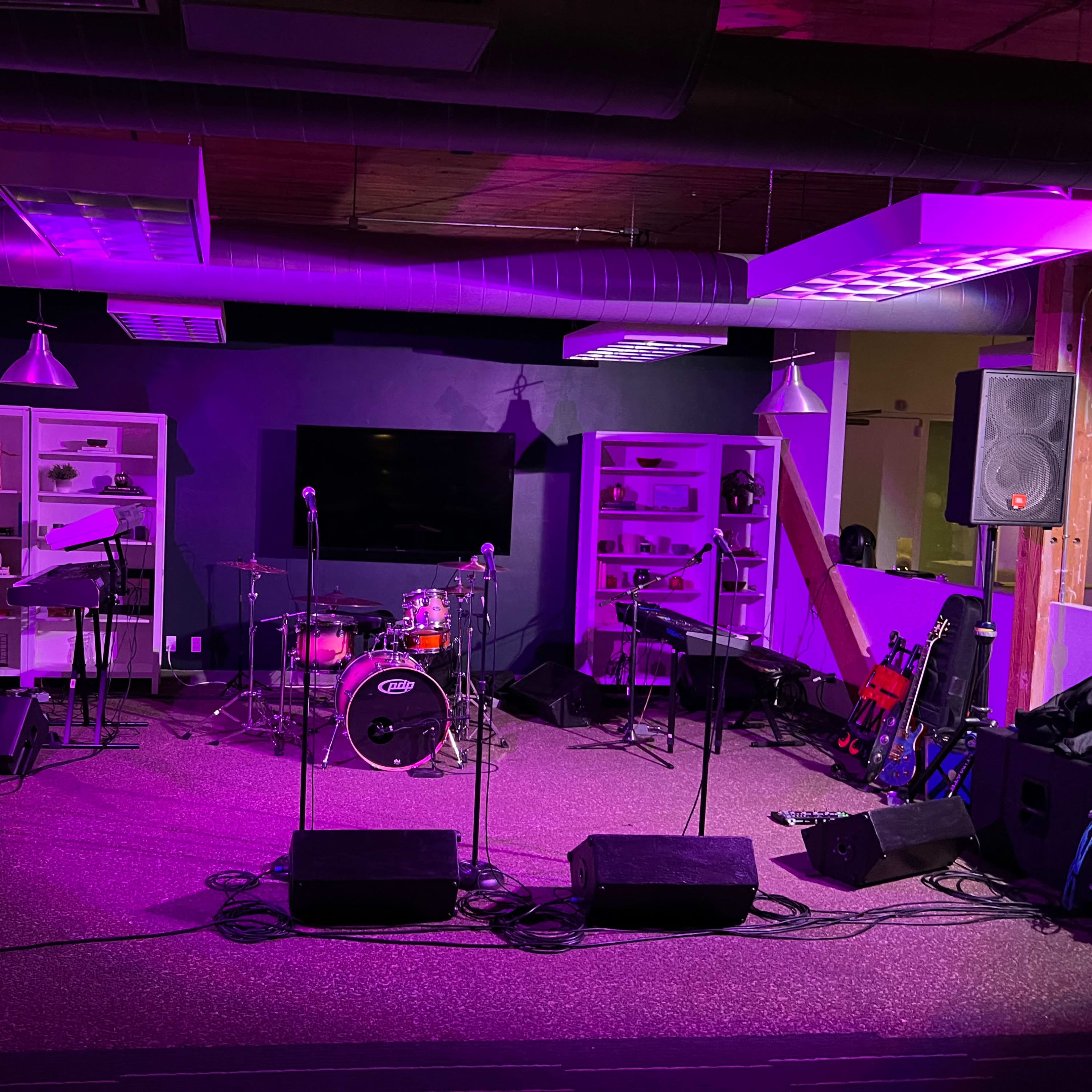 A small music stage setup with instruments, microphones, and sound equipment against a backdrop of shelves and a large screen.