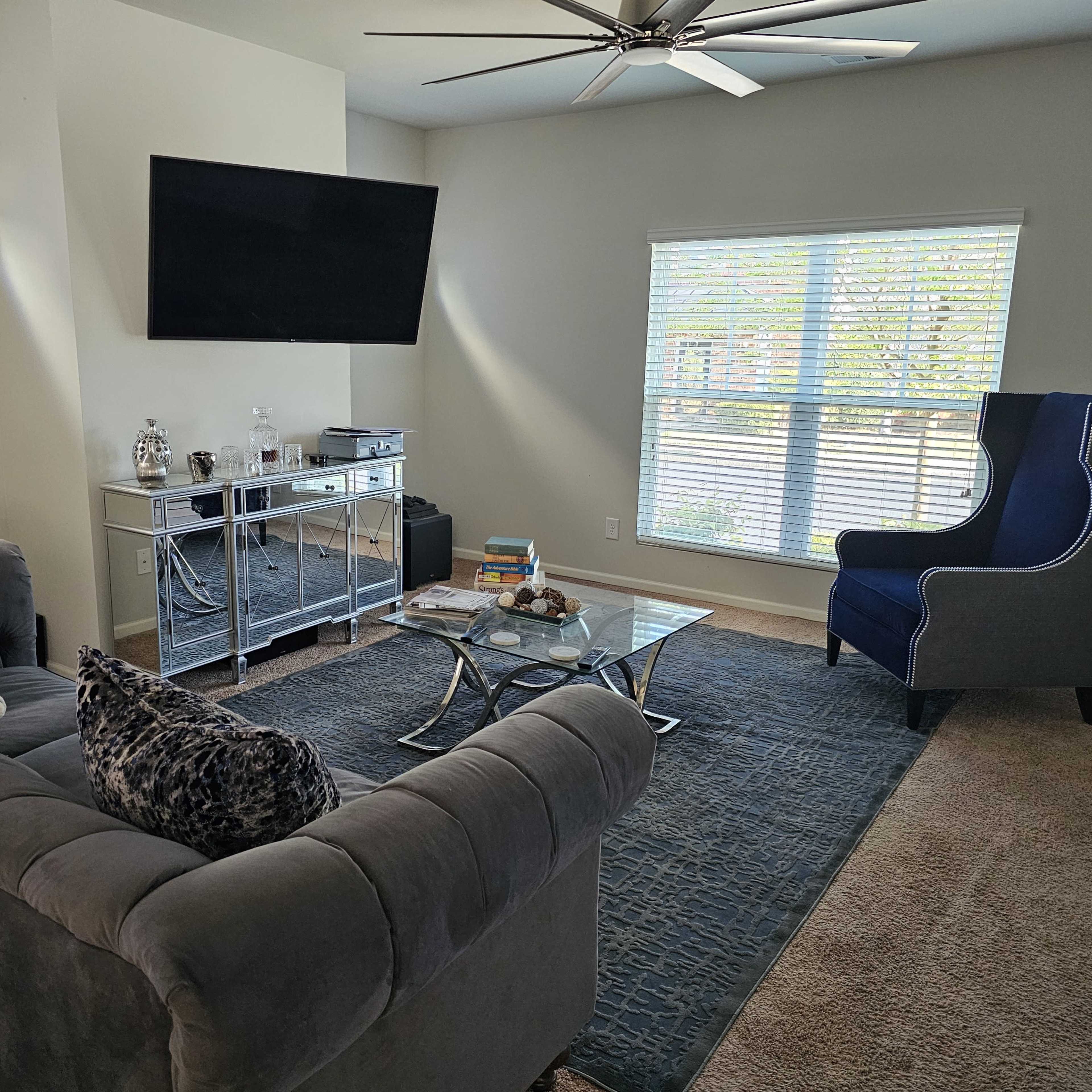 A living room with a gray sectional sofa, a glass coffee table, a large TV mounted on the wall, and a blue accent chair next to a window with blinds.