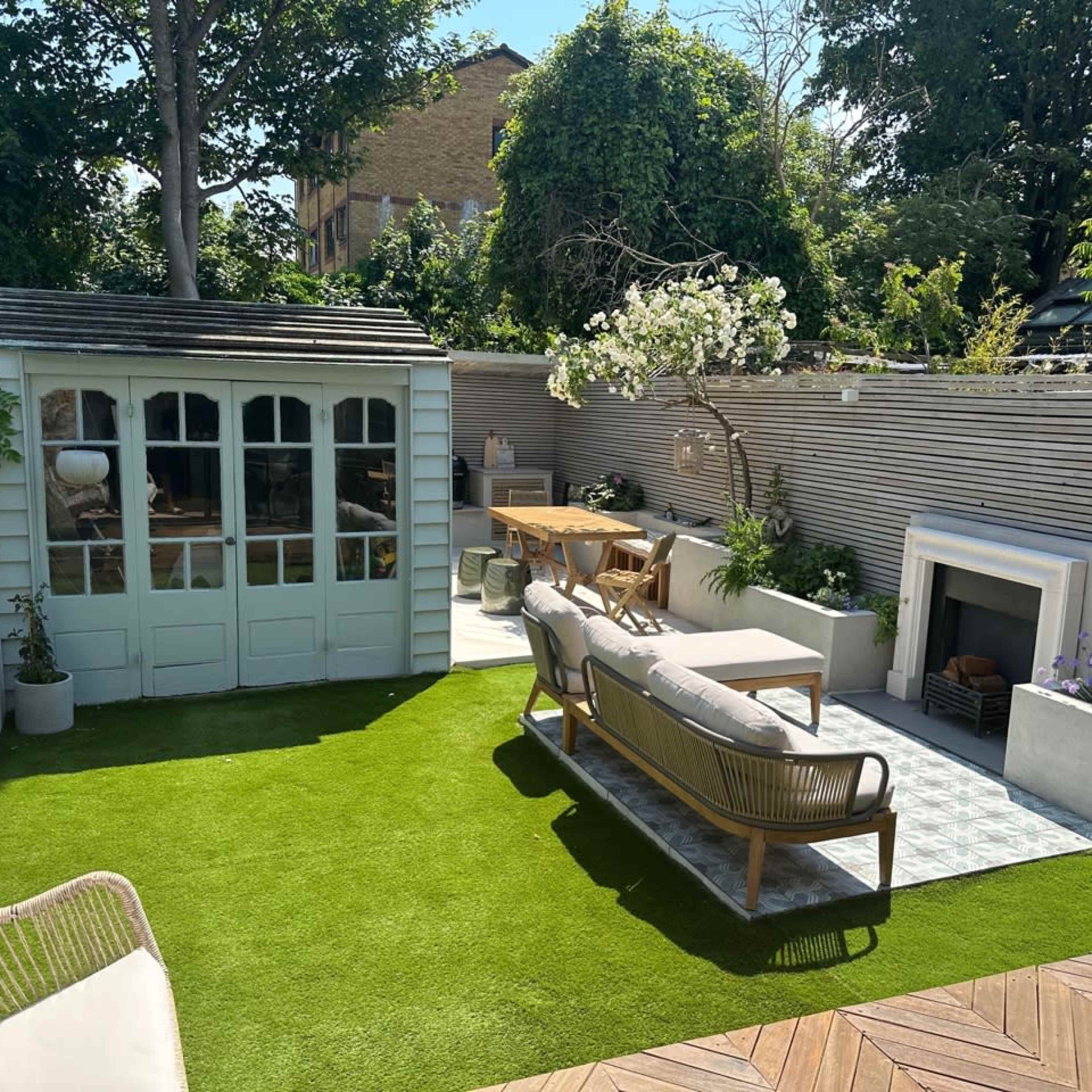 A landscaped garden featuring a lawn, outdoor seating area, a wooden dining table, and a small shed surrounded by greenery and a patio.