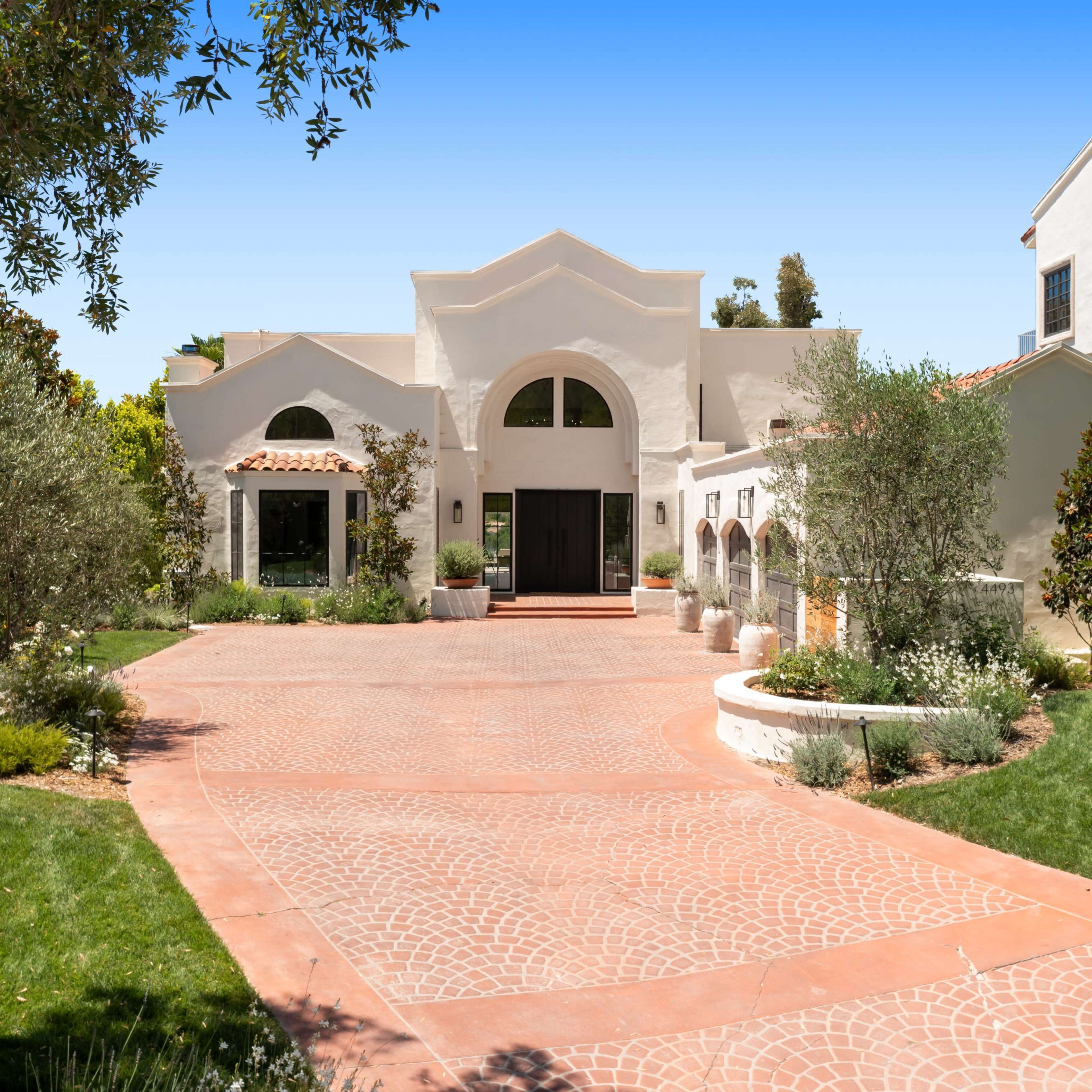 A large, white stucco house with a red brick driveway and landscaped greenery surrounding it.